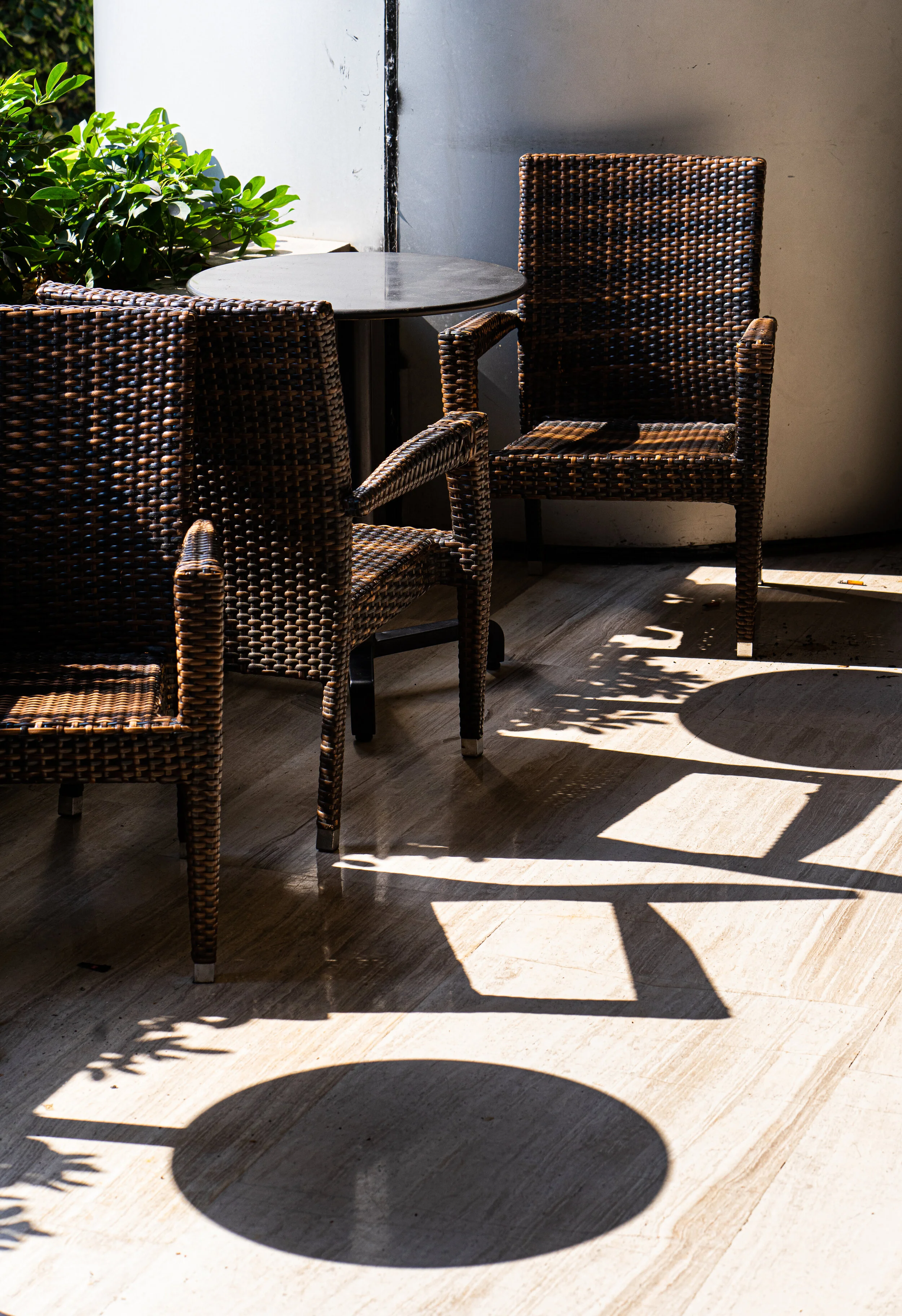 Sunlight casts shadows of two wicker chairs and a small round table on a wooden floor, with some greenery and a white wall in the background.