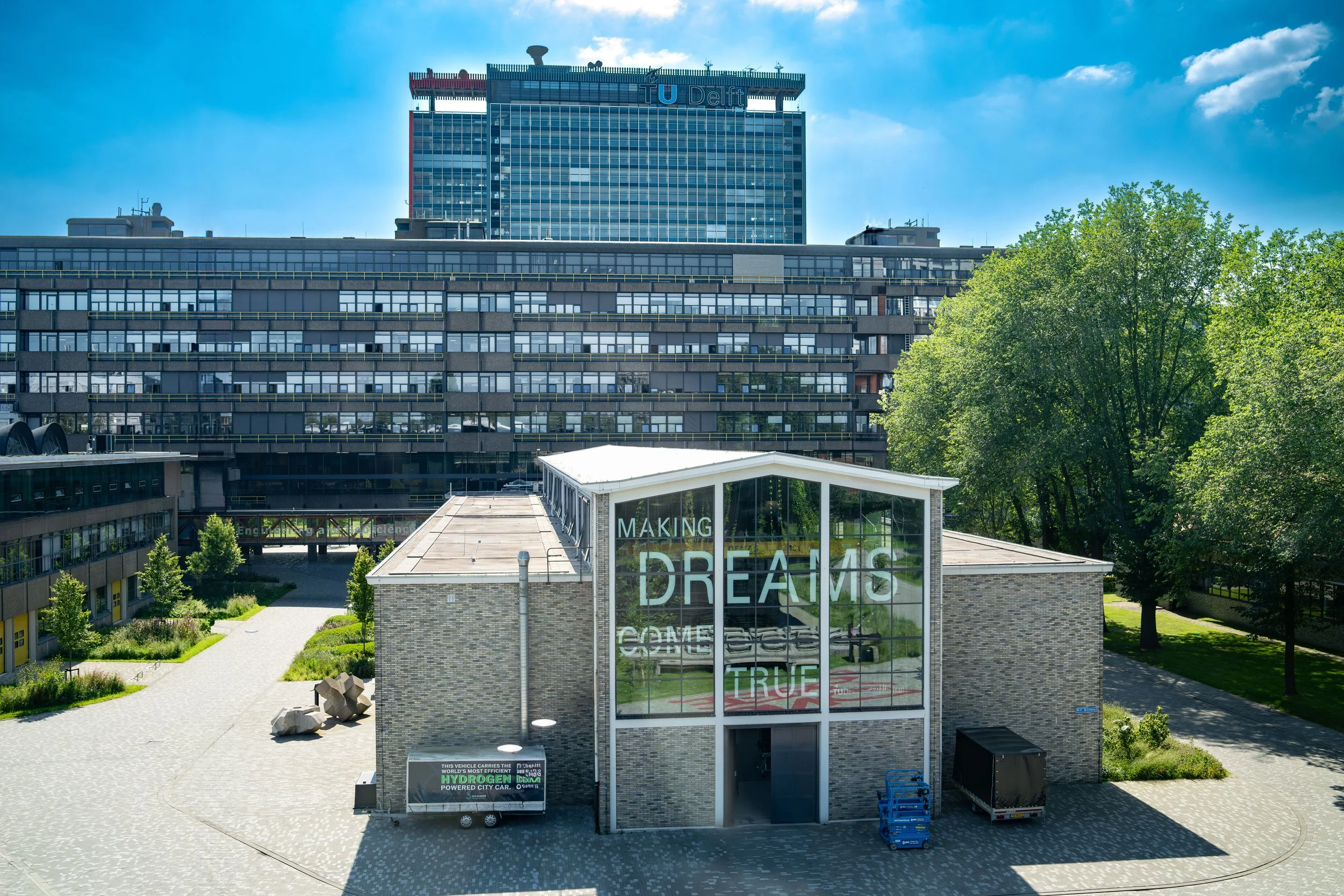 Modern building with glass facade displaying the words 'Making Dreams Come True' in large letters, surrounded by trees and city infrastructure under a partly cloudy sky.
