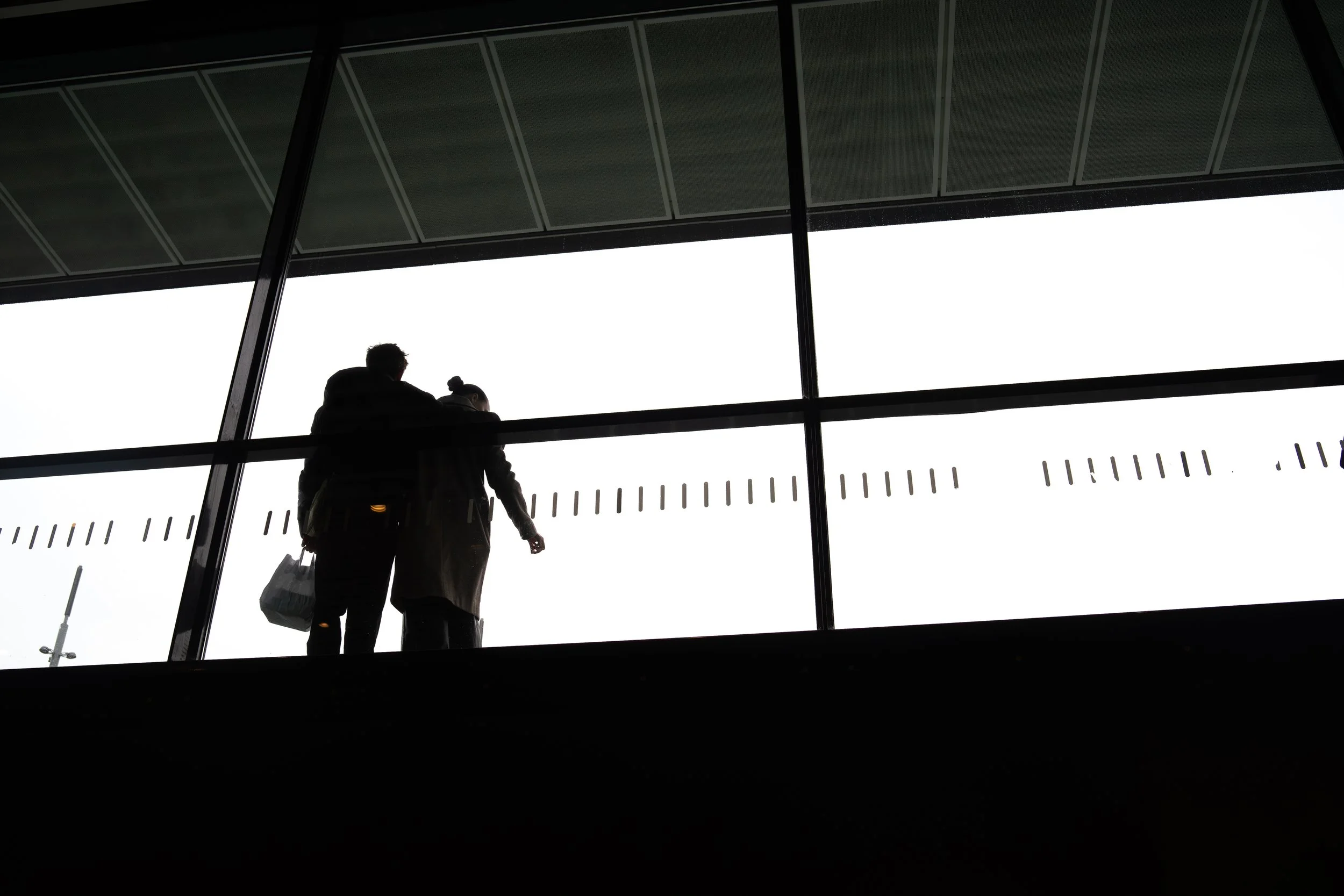 Silhouettes of two people standing on a bridge or balcony, looking out through large window panels at an overcast or bright sky outside.