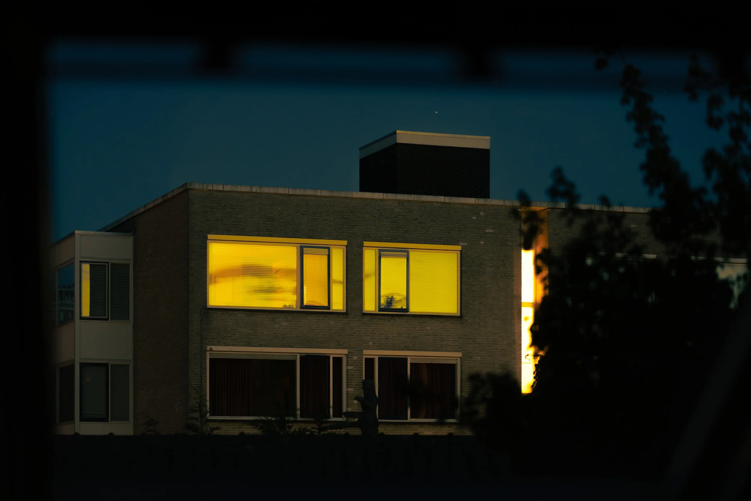 A modern multi-story building at night with yellow-lit windows and dark exterior walls, seen through a window from outside.