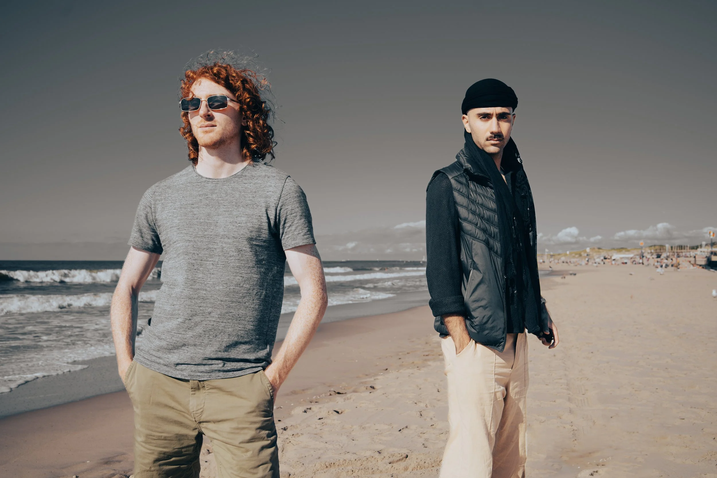 Two men standing on a sandy beach with the ocean in the background, one with curly red hair and sunglasses, the other with a turban and mustache.