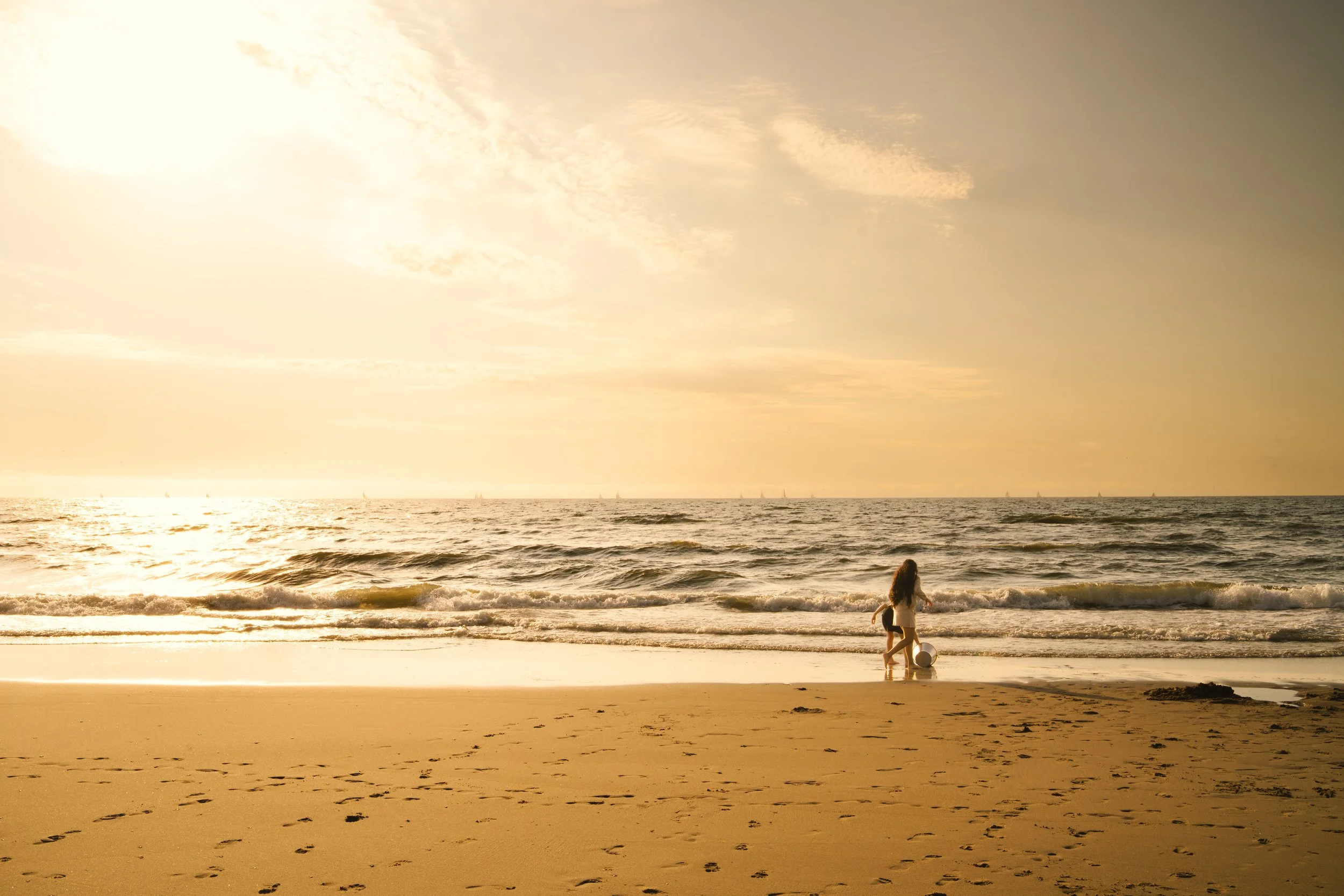A person and a dog on a sandy beach at sunset, with the ocean and sailing boats in the background.