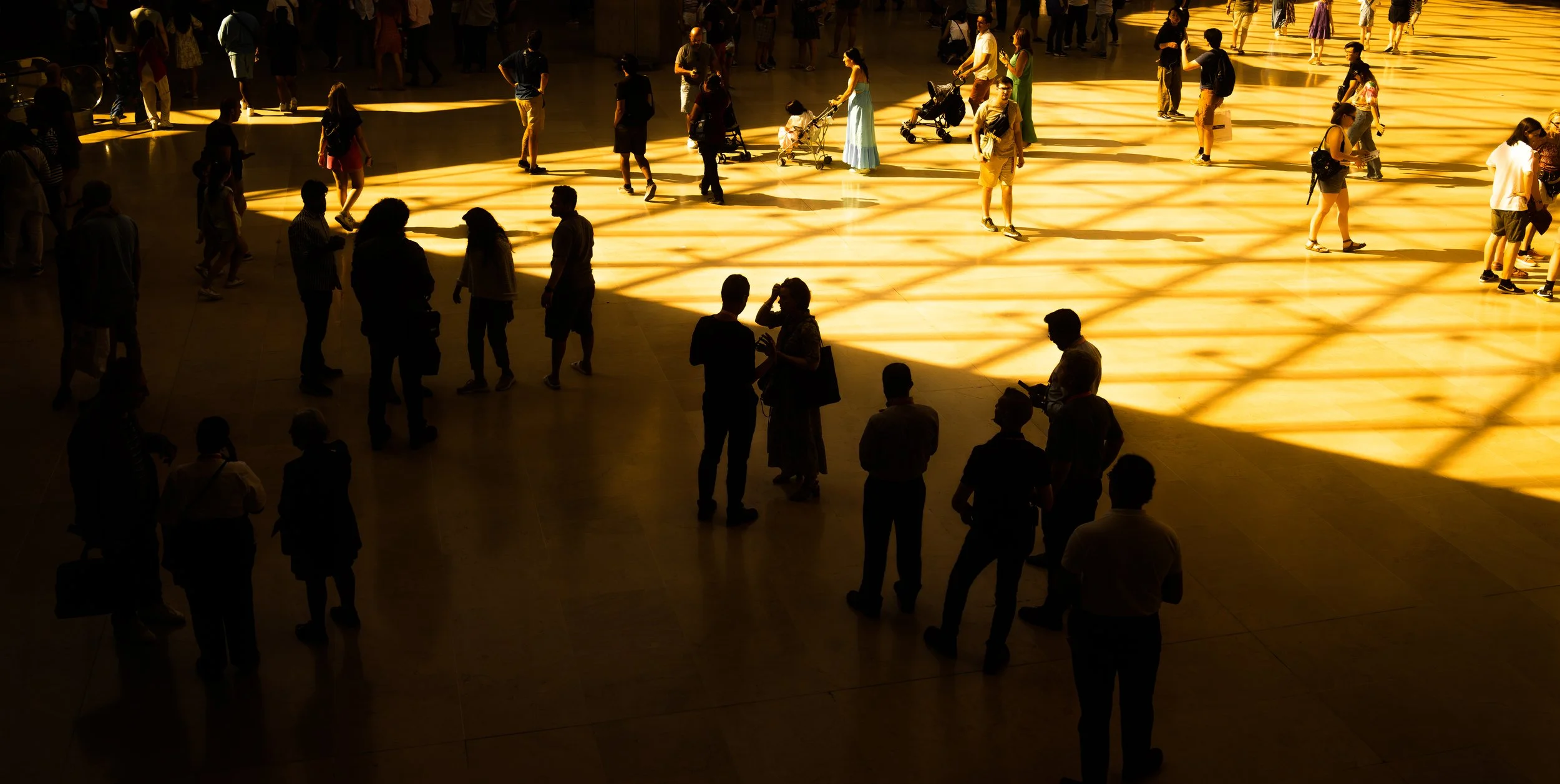 People gathering and walking inside a large, sunlit hall with patterned shadows on the floor.