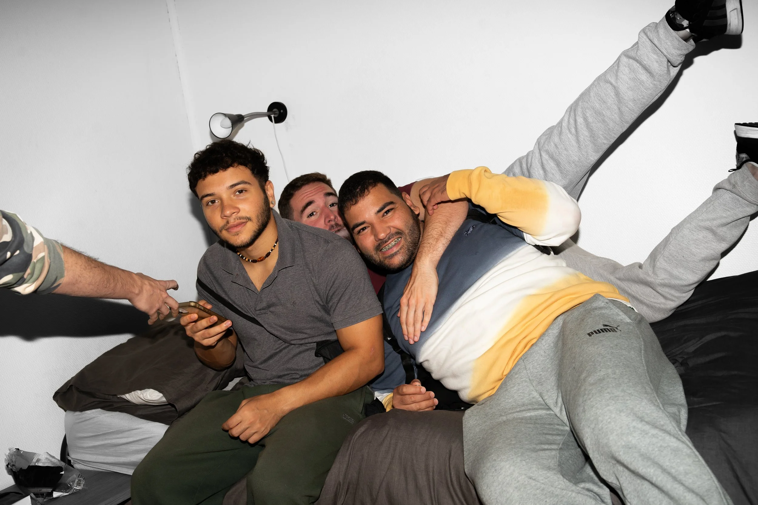 Four young men sitting on a bed, smiling, with one of them using a cellphone, and another has arm around a friend in a casual indoor setting.