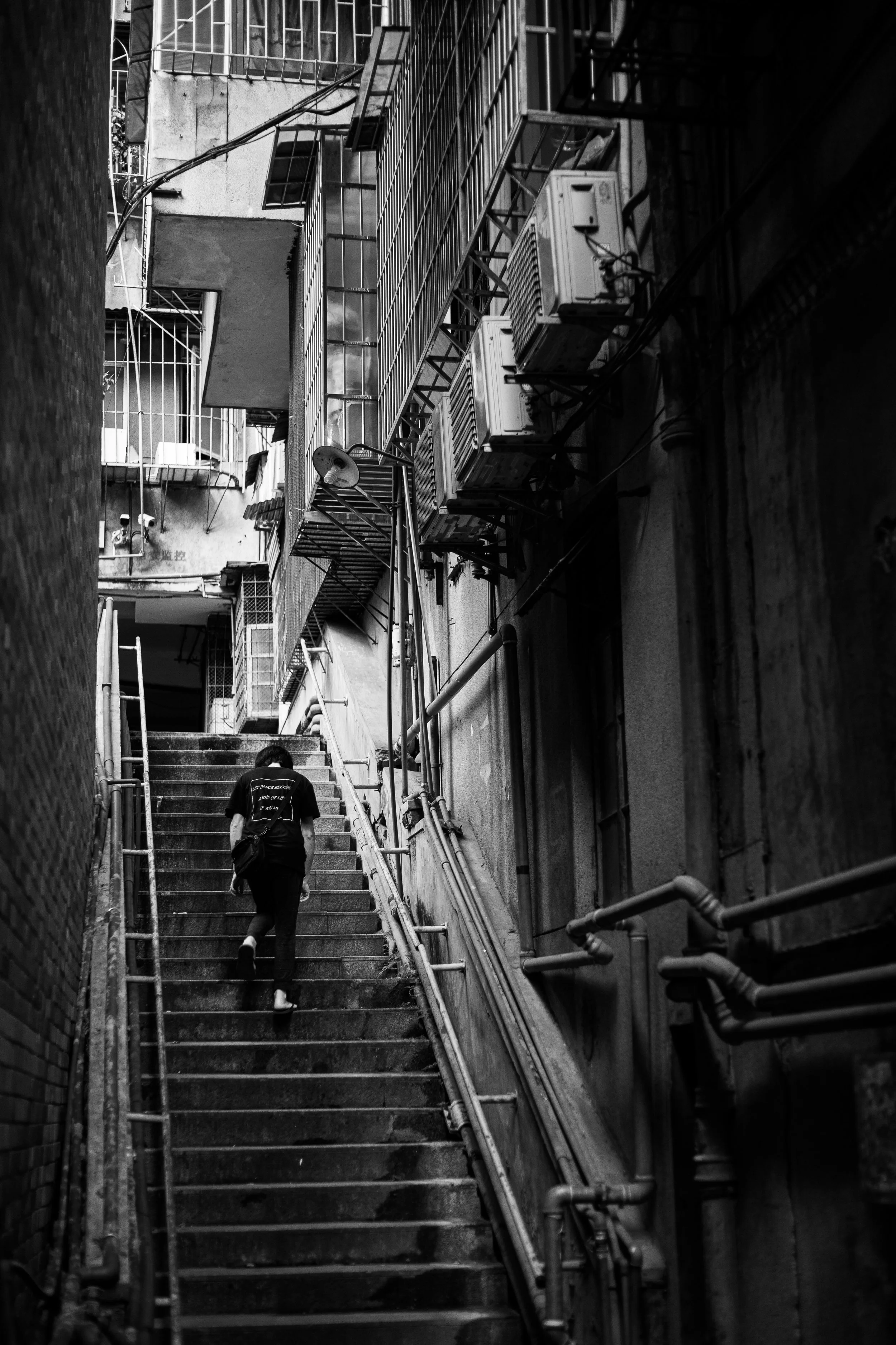 A person wearing a black shirt and carrying a bag is ascending a narrow outdoor staircase in an urban setting. The staircase is surrounded by tall buildings with visible air conditioning units and pipes on the walls. The photo is in black and white.