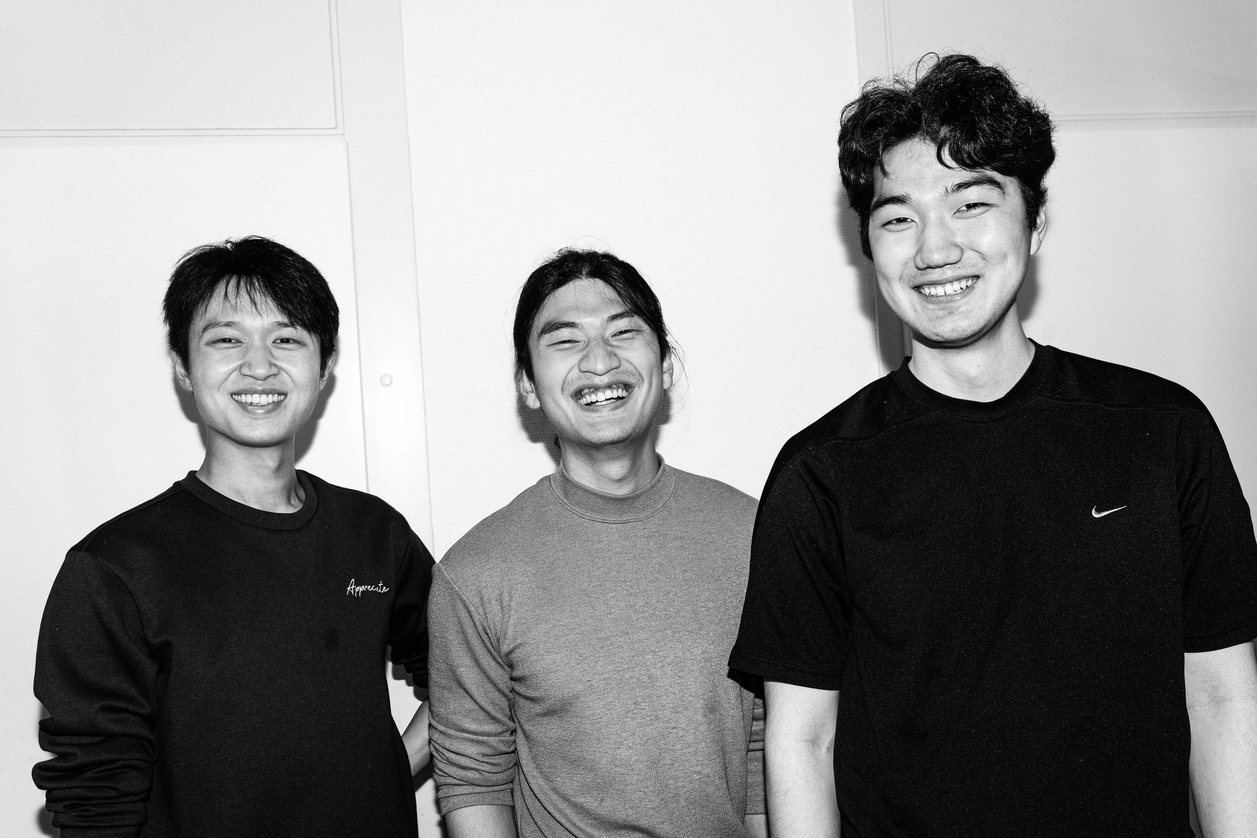 Three young men smiling and laughing together, standing indoors against a plain wall.