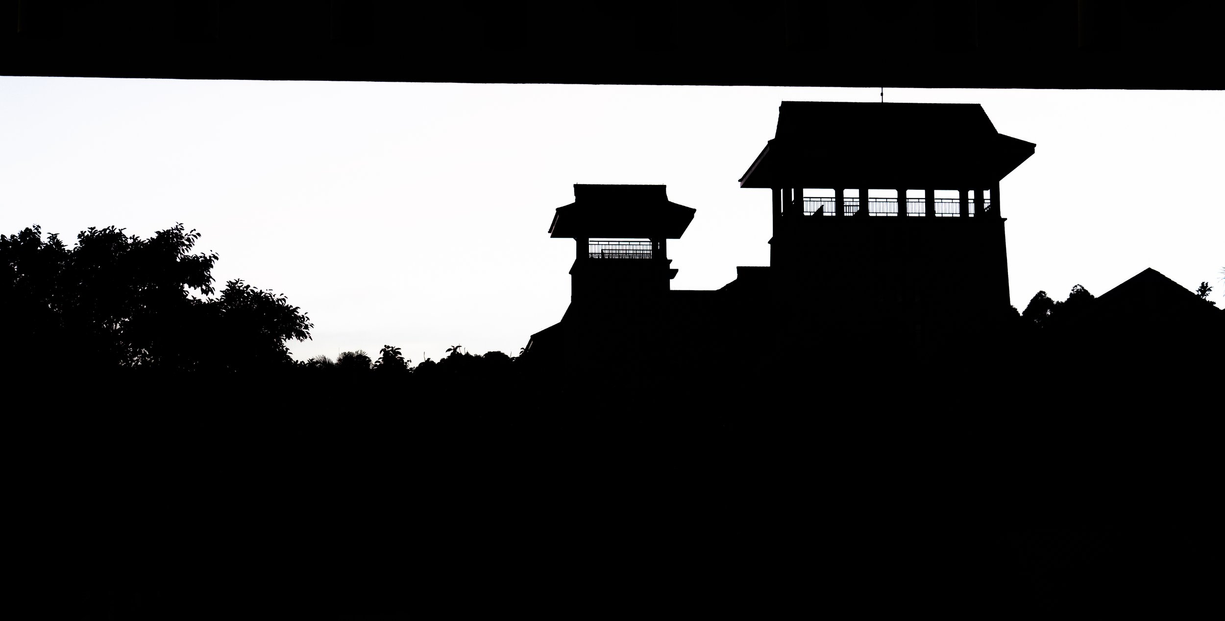 Silhouette of a castle or fortress with watchtowers and trees in the foreground, seen against a bright sky.