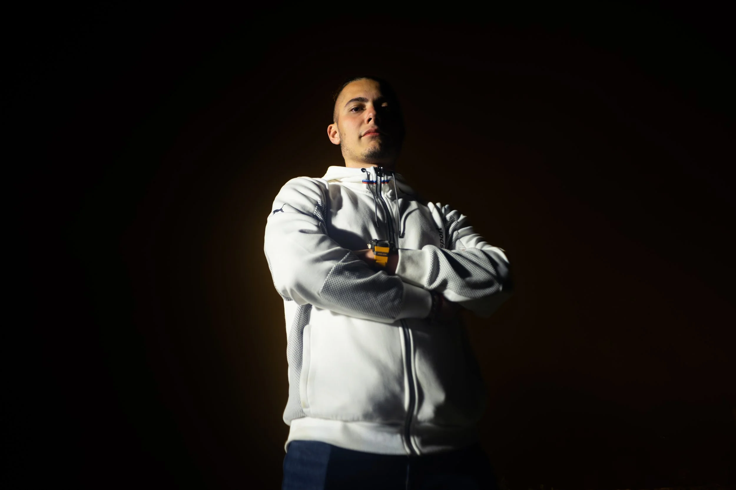 Young man with arms crossed, wearing a white athletic jacket, standing confidently with a dark background and subtle lighting highlighting his upper body.