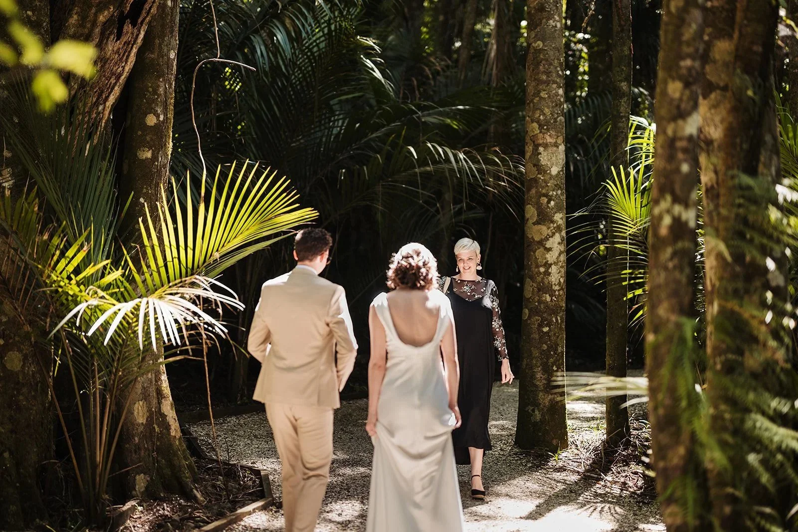 A couple in wedding attire walking through a lush, shaded forest with an officiant faces them, holding a wedding ceremony.