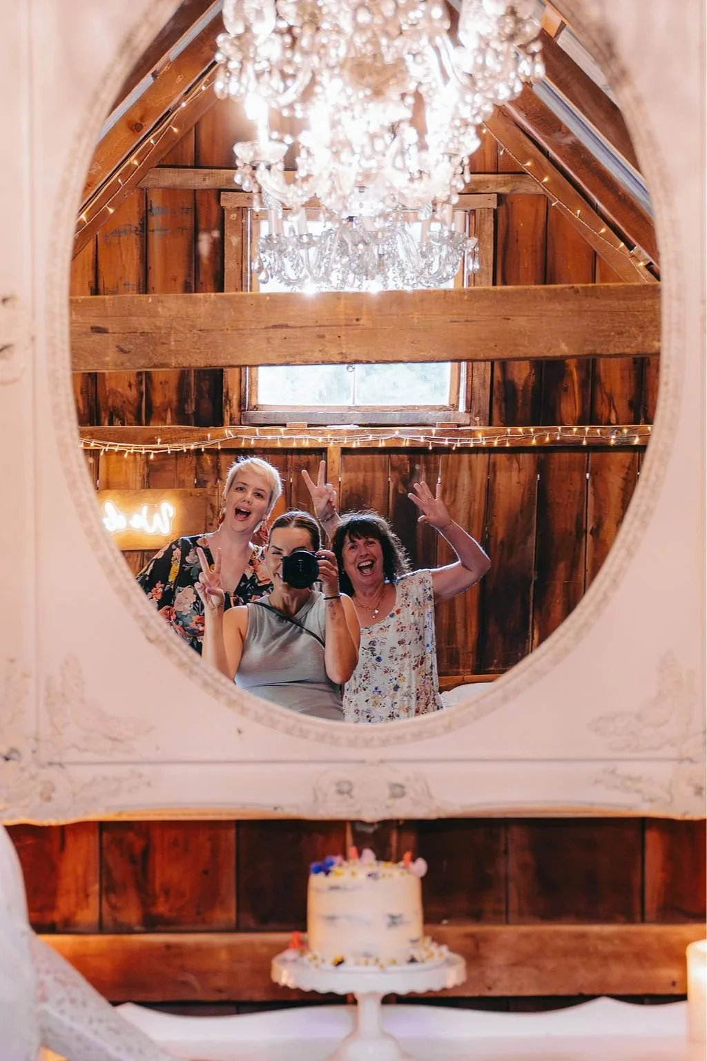 Three women taking a selfie through a rustic mirror in a wooden room decorated with string lights and a chandelier, with a cake on the table in front of them.