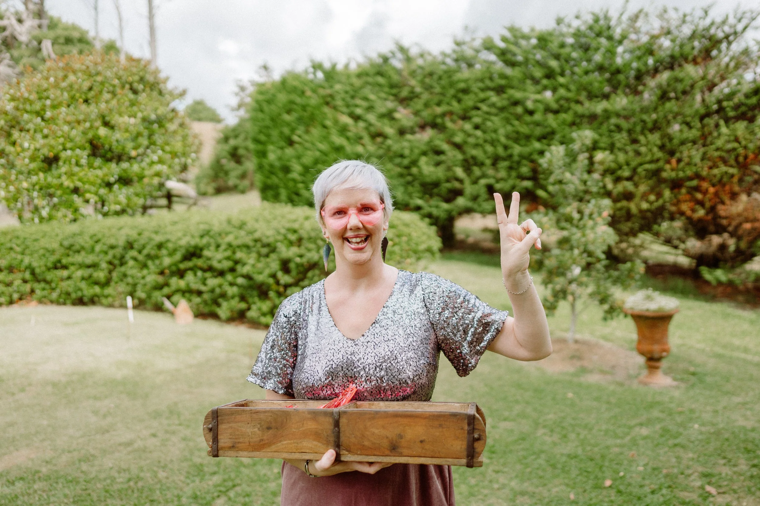 A woman with short gray hair, pink glasses, and earrings outdoors making a peace sign with her right hand, holding a wooden basket with red some produce, standing in front of green trees and bushes.