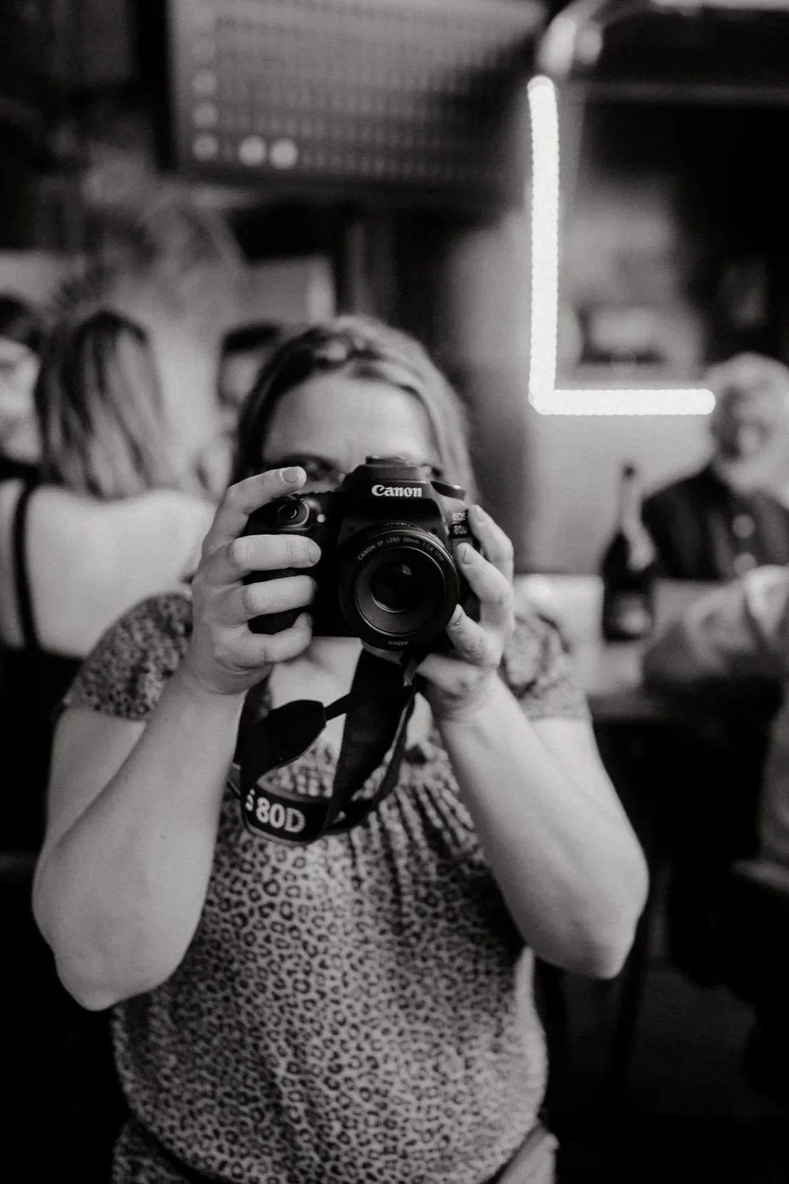 A woman taking a photo with a Canon camera in a black-and-white setting, with two other women in the background and a neon light in the shape of an L in the upper right corner.