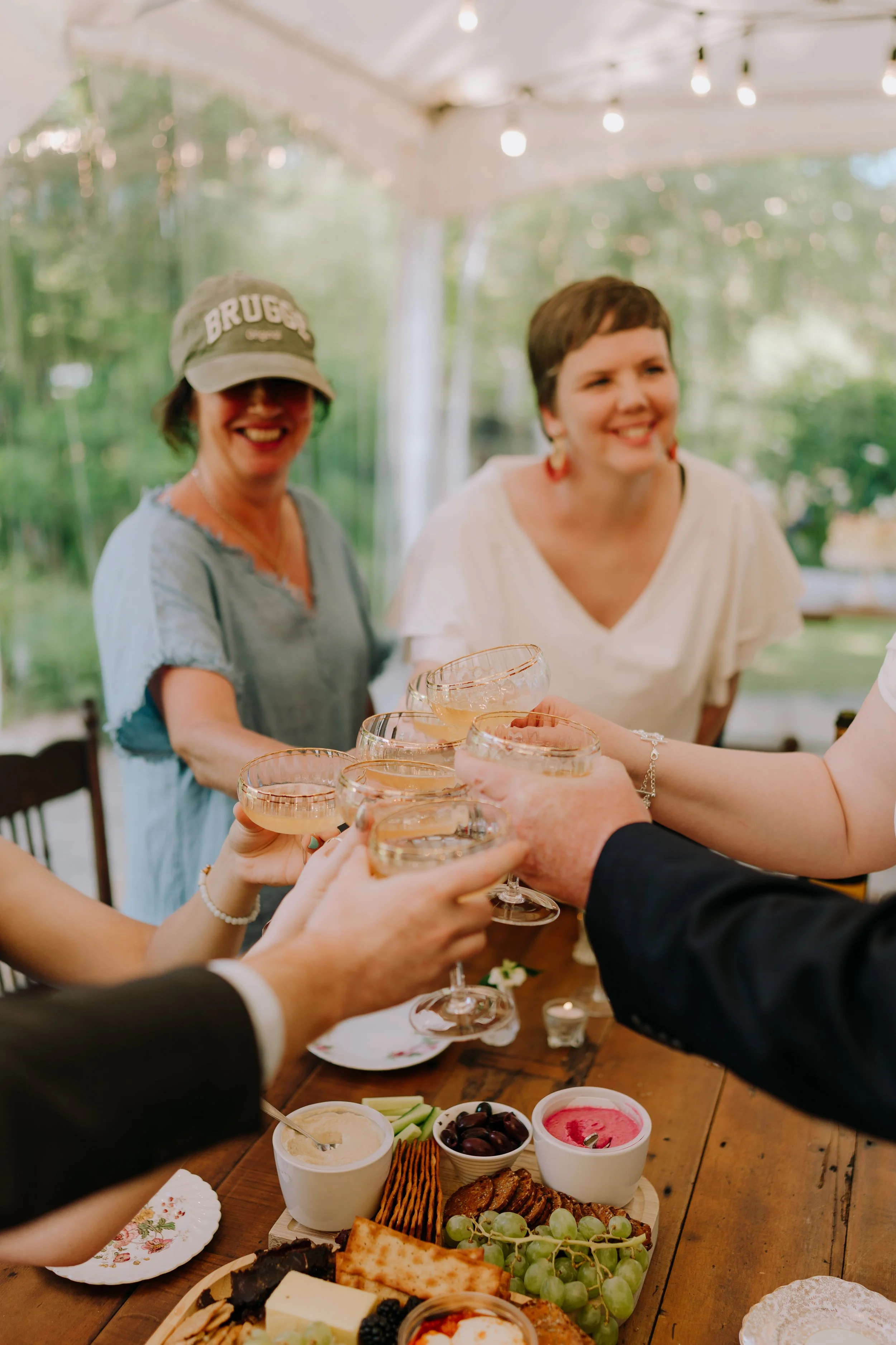 People at a celebration toast with champagne glasses at a gathering with food and drinks on a wooden table. The background is decorated with string lights and greenery.