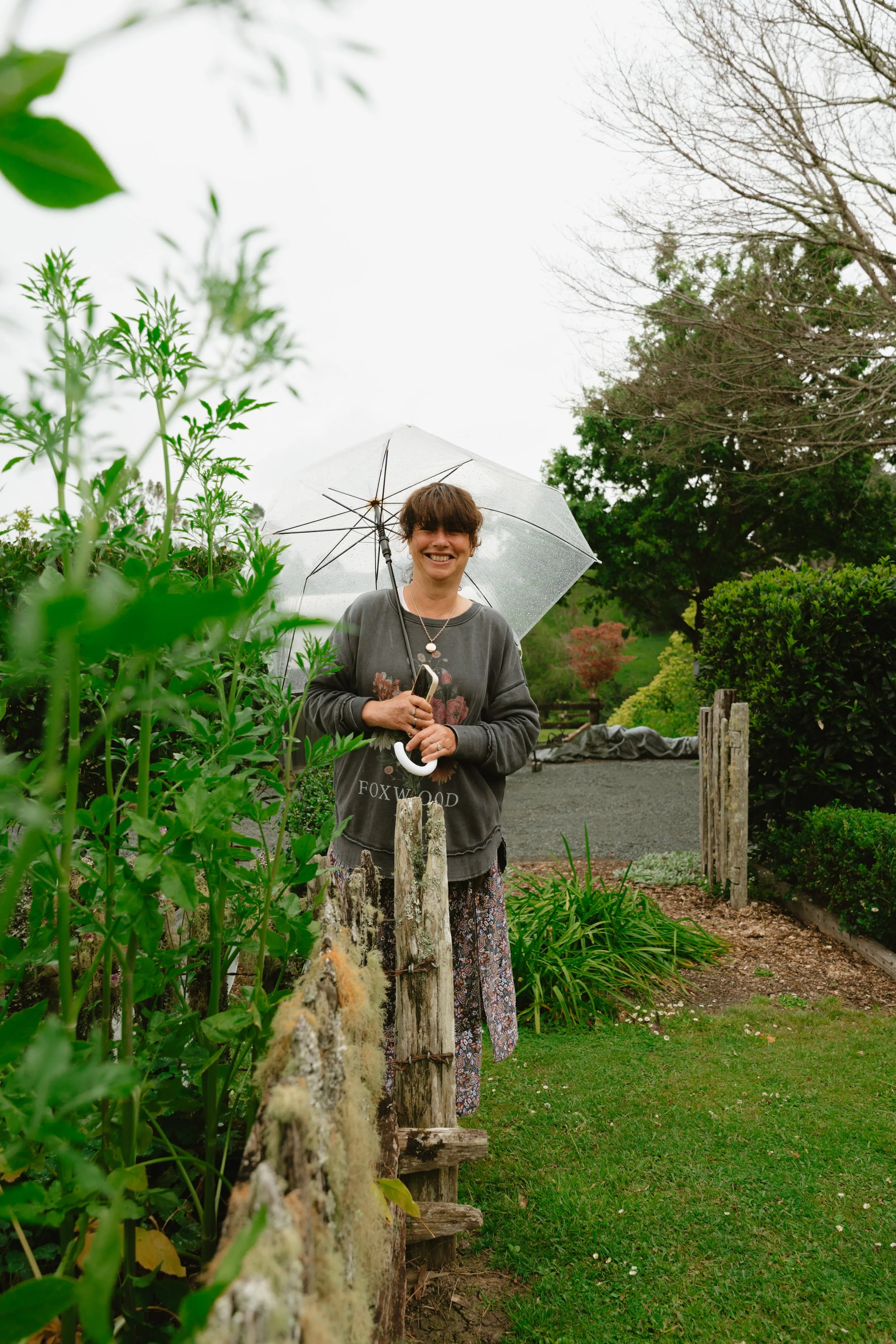 Smiling woman holding a clear umbrella stands in a garden surrounded by lush green plants and bushes during an overcast day.