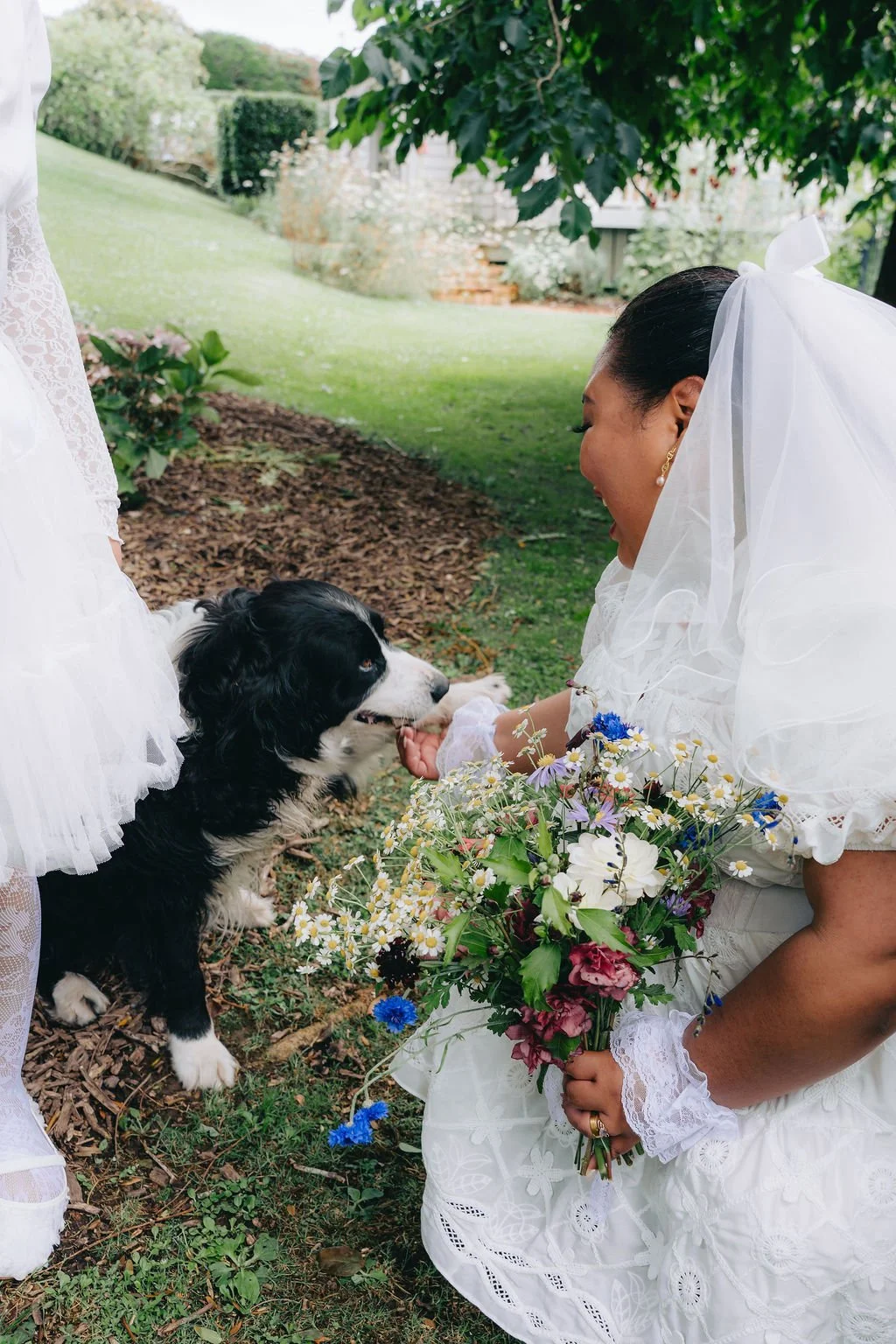 A woman in a white dress and veil is smiling as she gently interacts with a black and white dog during a garden wedding. She holds a colorful bouquet of flowers, and the outdoor setting includes lush greenery and flowering plants.