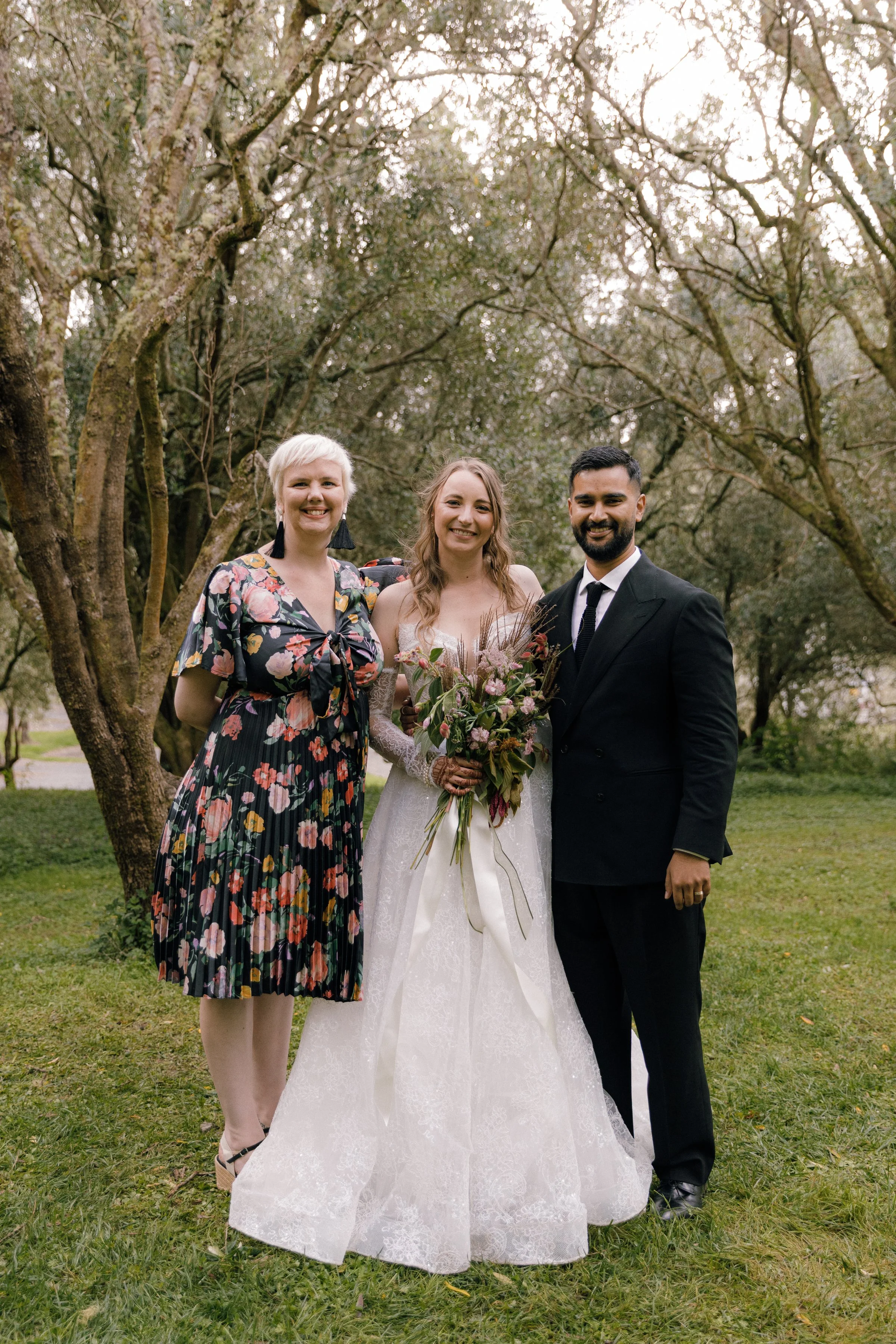 Three people standing outdoors among trees, with a woman in a wedding dress holding a bouquet, flanked by a woman in a floral dress and a man in a suit, all smiling.