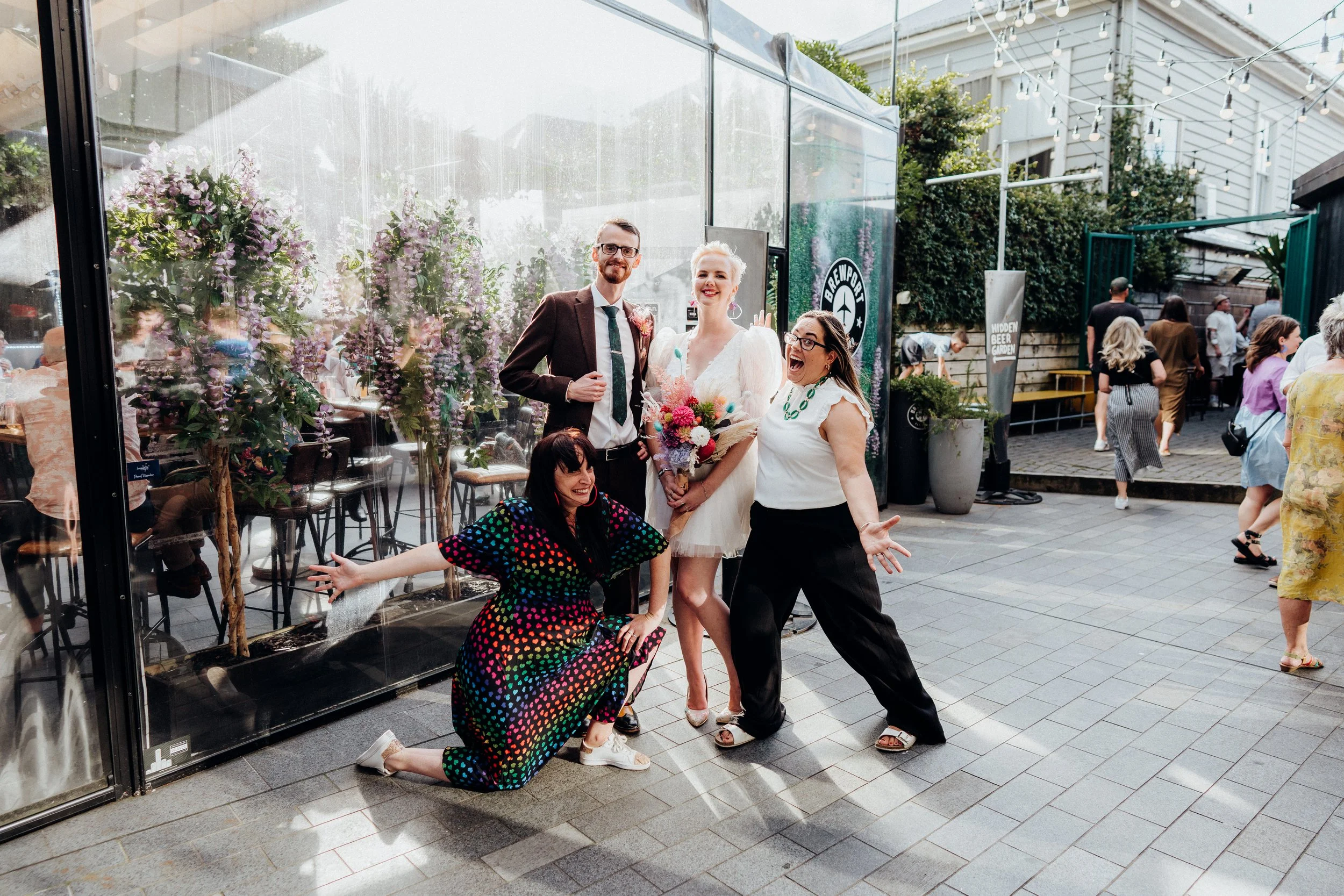 Group of four people smiling and posing outside a glass-walled venue with purple flowering trees inside, during daytime, with other people walking by in the background.