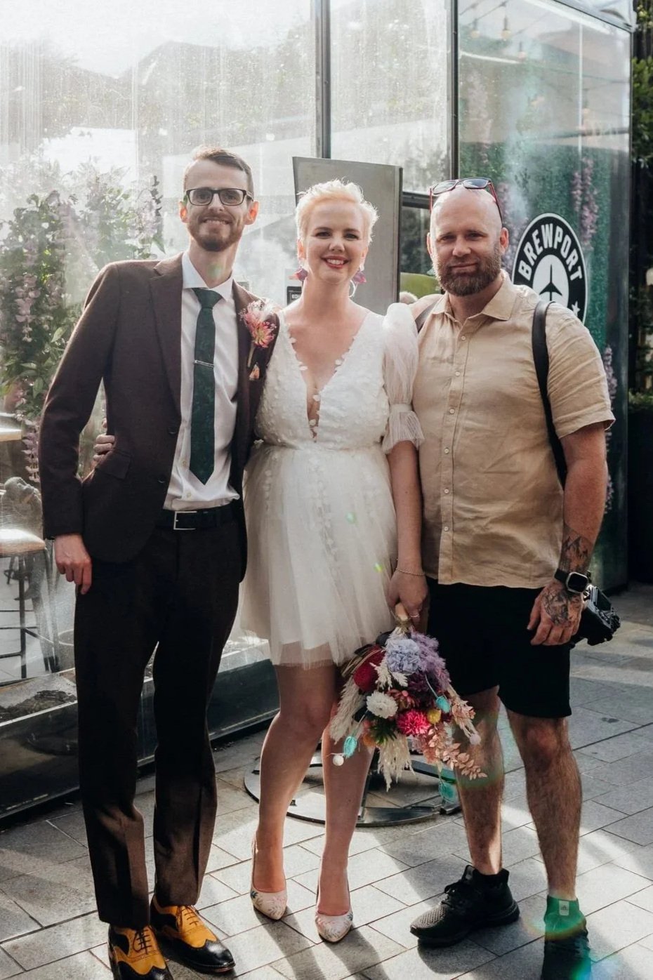 Three people posing together inside a garden. The person in the middle is wearing a white dress and holding a bouquet, indicating a wedding. The person on the left is in a suit with a tie, and the person on the right is casually dressed in a beige shirt and shorts with tattoos