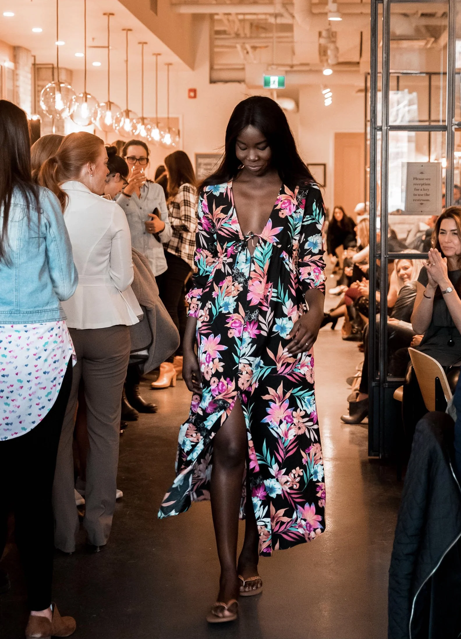A woman walking on a runway indoors, wearing a floral dress with a deep neckline. She is surrounded by an audience observing her, in a well-lit room with pendant lights and seated guests.