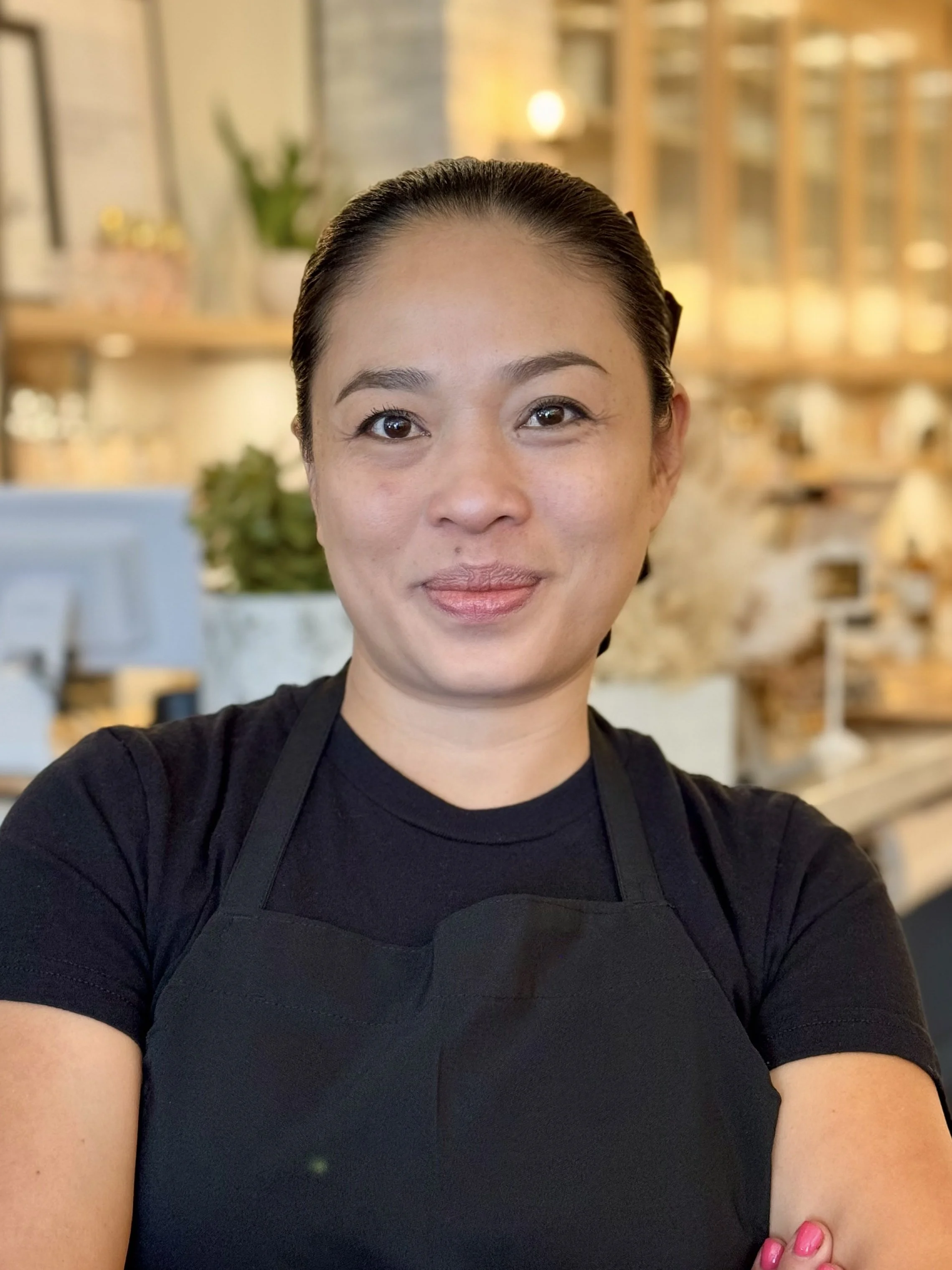 A woman with dark hair pulled back, wearing a black shirt and apron, smiling in a warmly lit coffee shop or cafe with wooden shelves and plants in the background.
