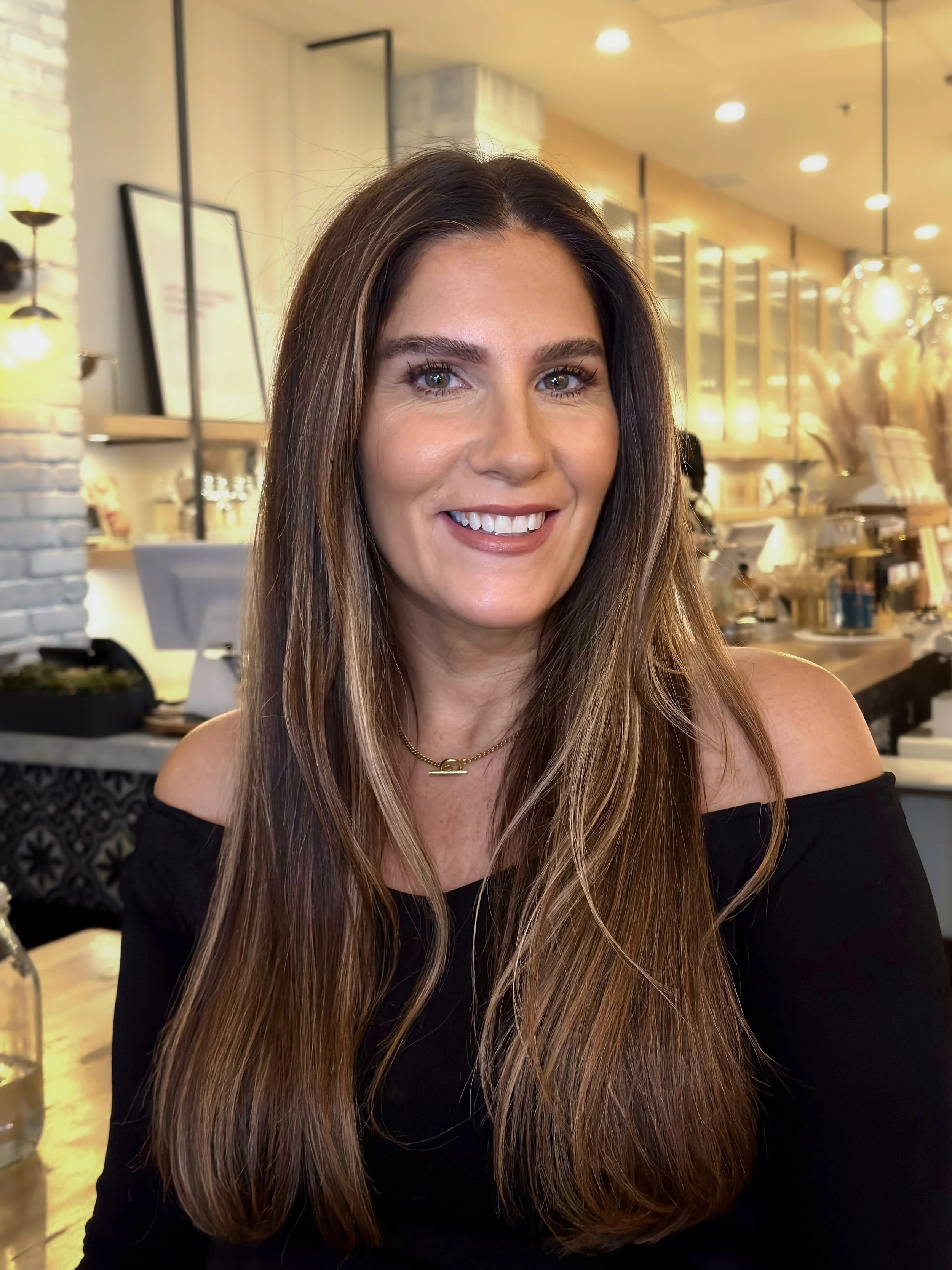 A smiling woman with long brown hair wearing a black off-the-shoulder top and a gold necklace, seated in a cozy café or restaurant with warm lighting and decorative items in the background.