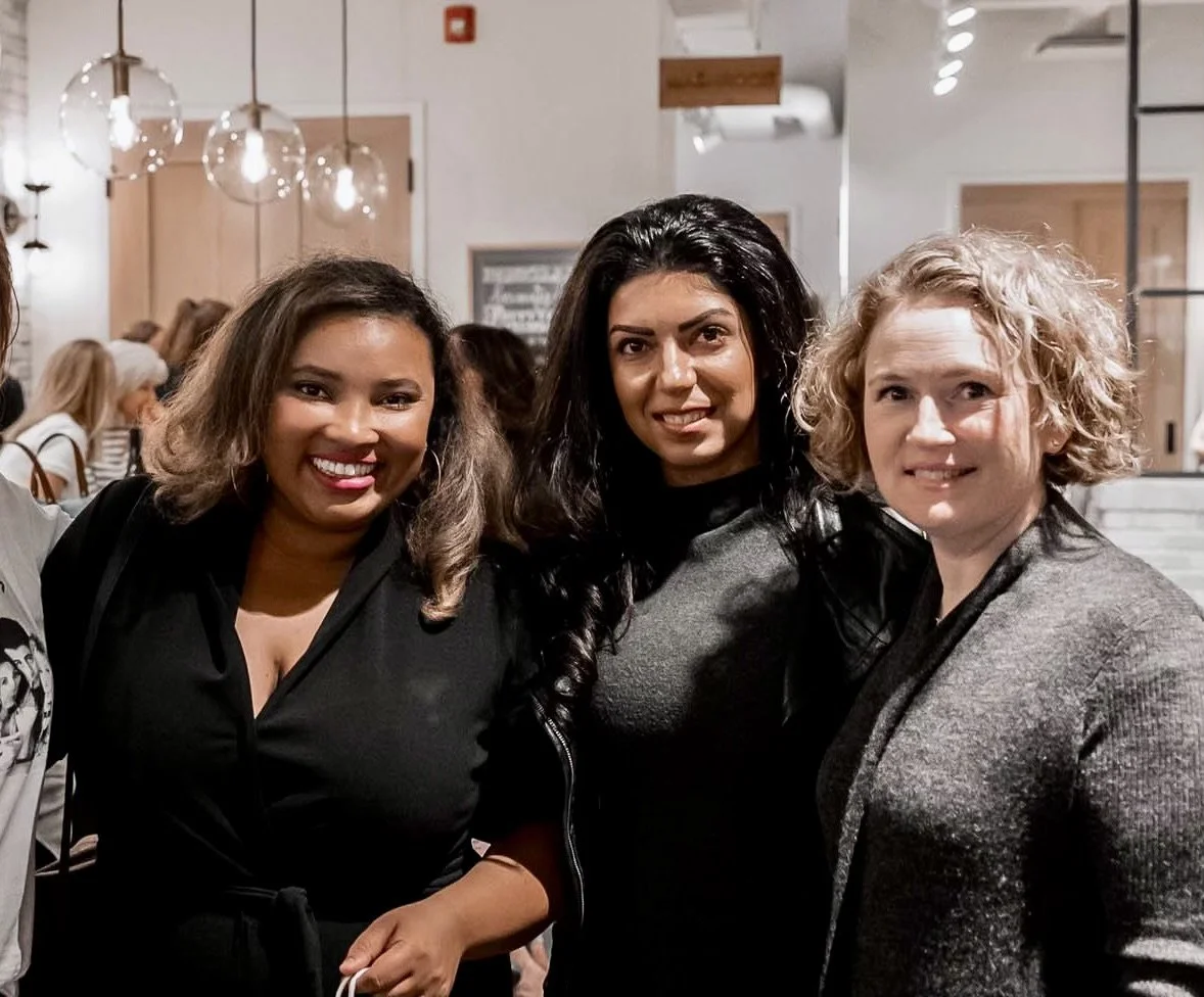 Three women smiling and posing together in a warmly lit indoor setting with modern decor.