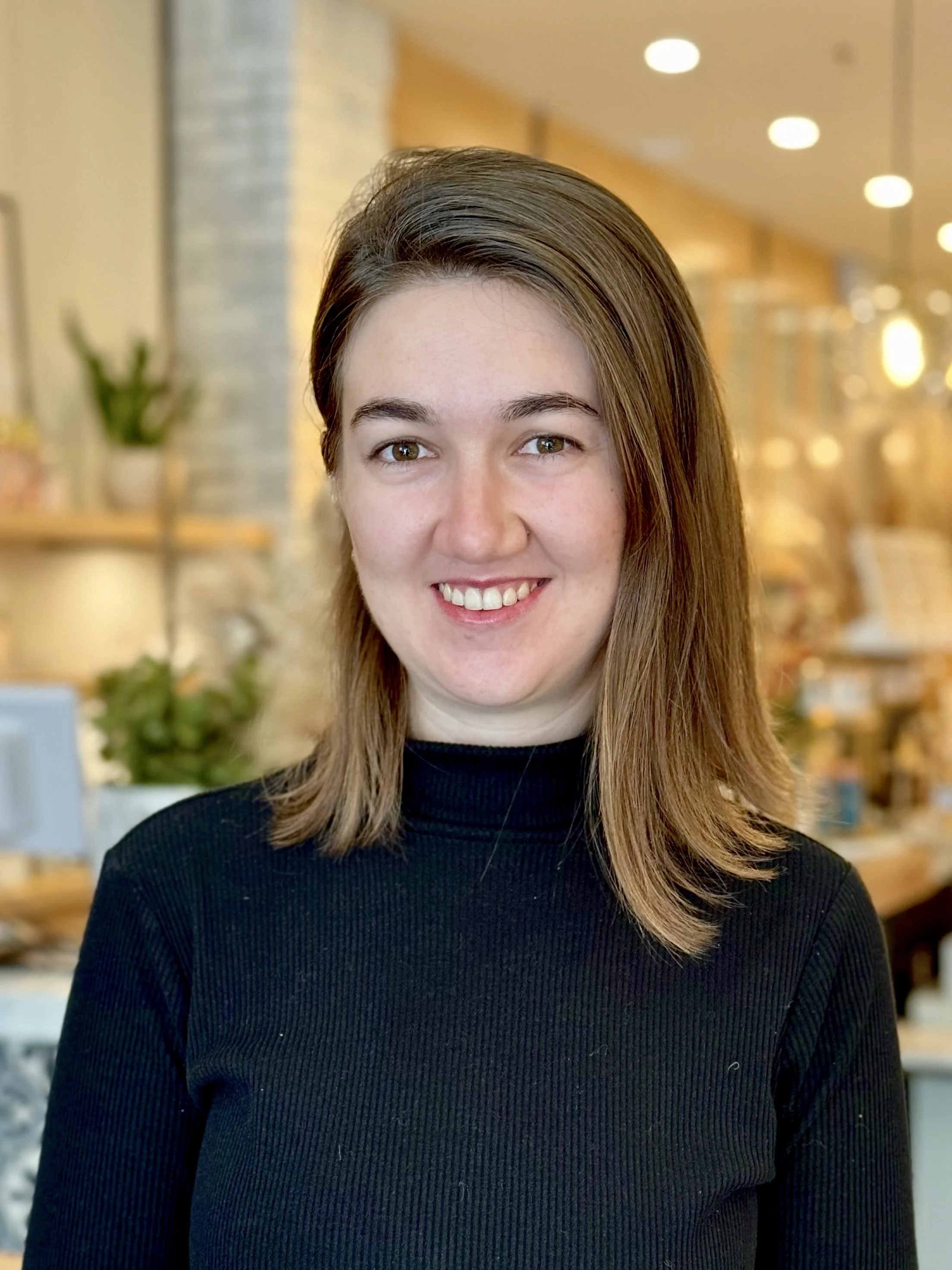 A young woman with shoulder-length brown hair smiling at the camera in a cozy indoor setting.
