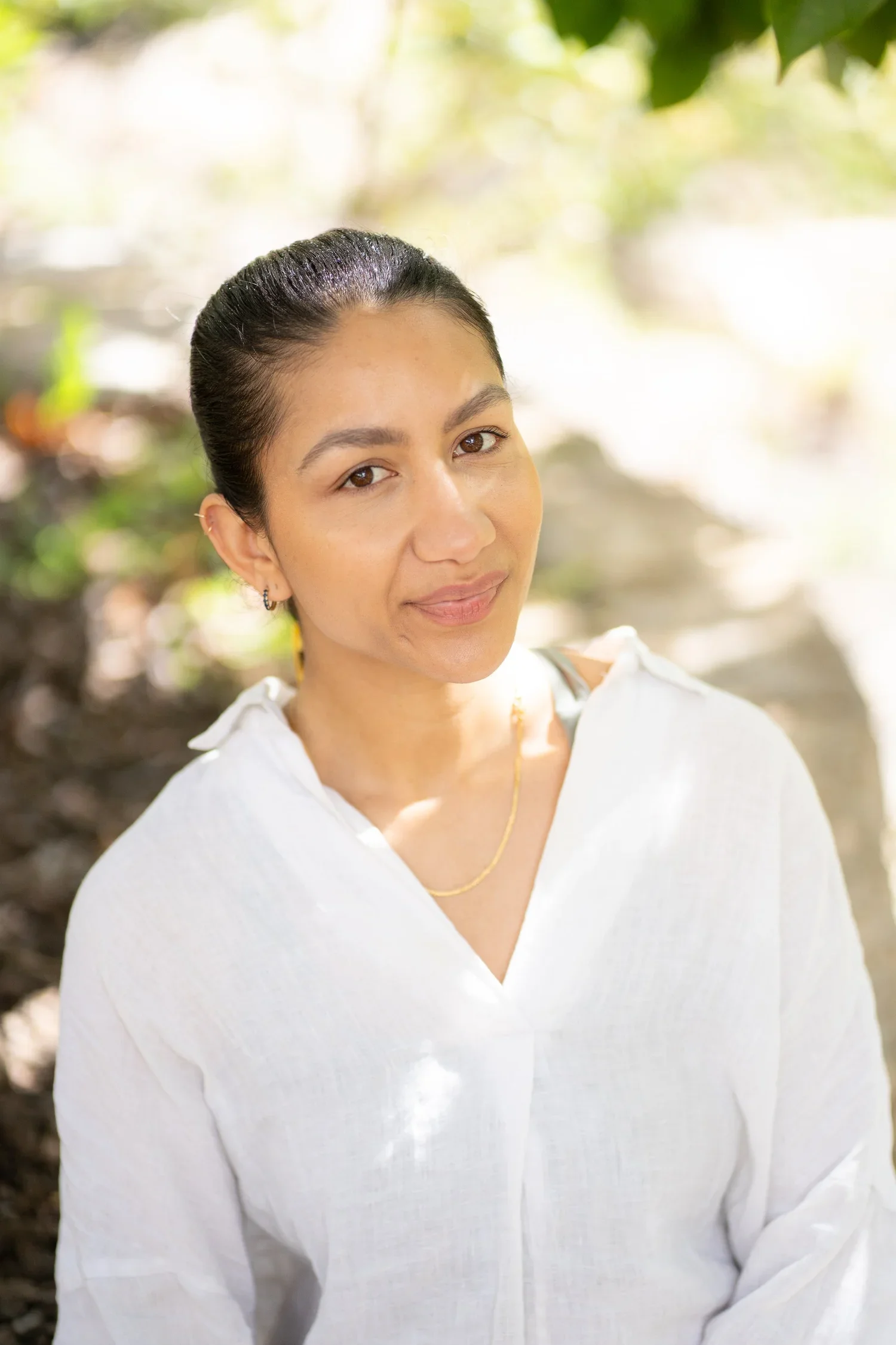 A woman with dark hair tied back, wearing a white blouse and a gold necklace, standing outdoors with greenery in the background.