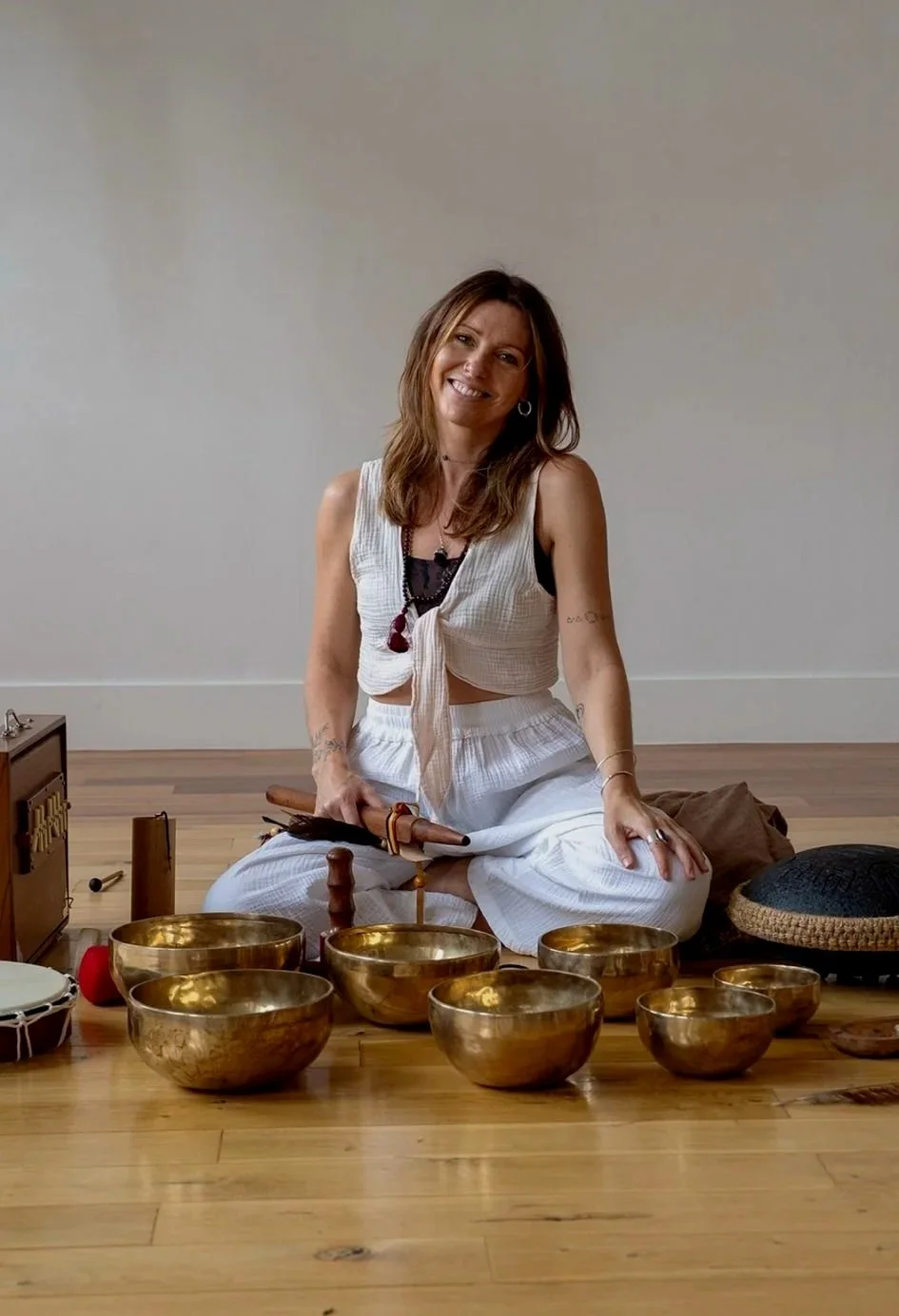 Woman sitting on the floor surrounded by singing bowls, holding a mallet, smiling indoors with a plain wall background.