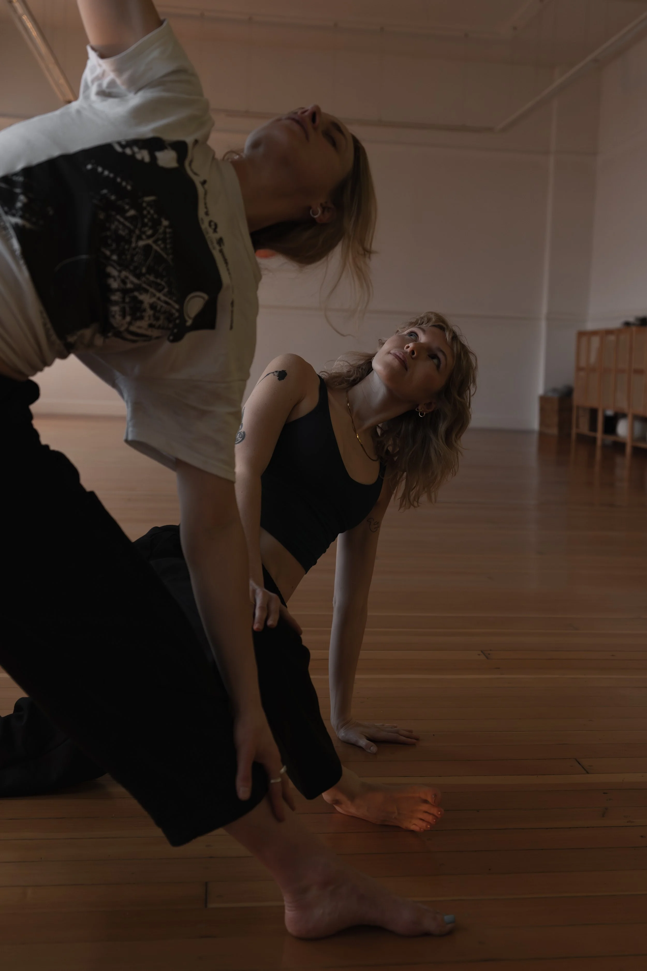 Two women practicing yoga in a wooden studio with one woman in a black tank top in a pose and the other leaning forward, wearing a graphic t-shirt, with natural light coming in.