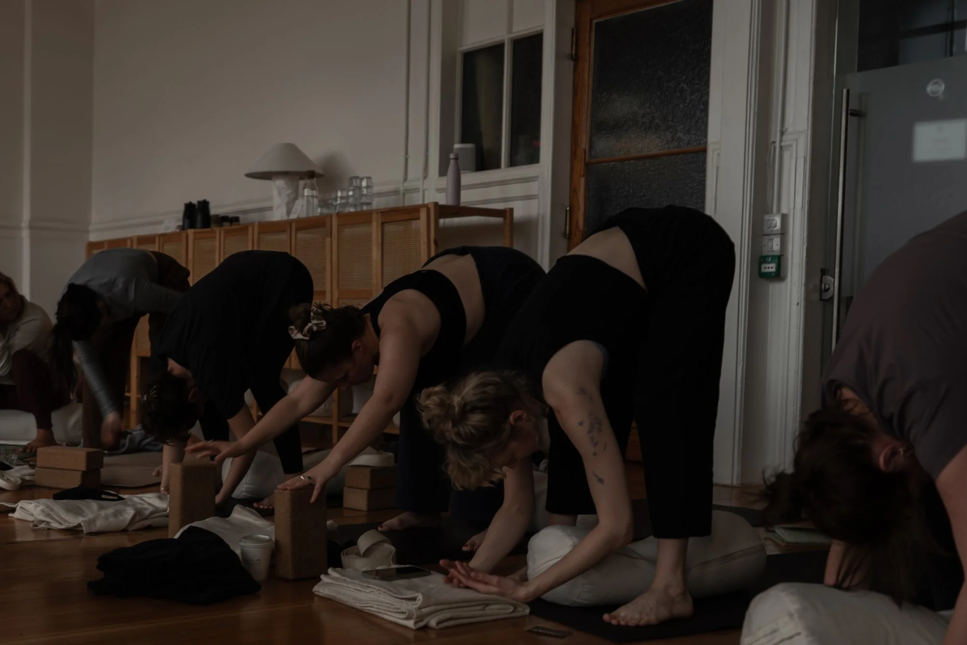 Group of people practicing yoga or stretching on a wooden floor in a room with white walls and large windows, with some objects like a lamp, bottles, and books on a wooden cabinet in the background.