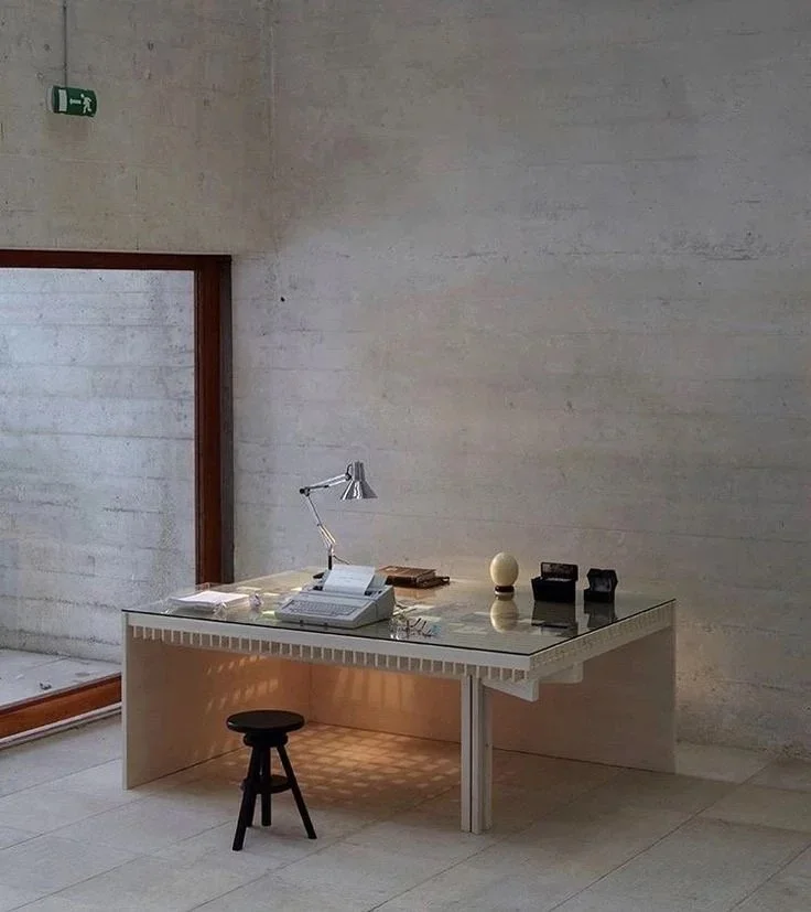 Minimalist office space with a white desk, black stool, and a large window, with a few items like a lamp, documents, and decorative objects on the desk.