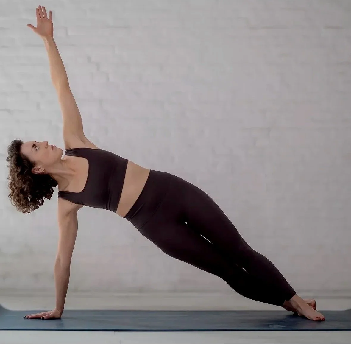 A woman practicing yoga indoors, performing a side plank pose with one arm extended upward and her body supported by her hands and feet on a yoga mat.