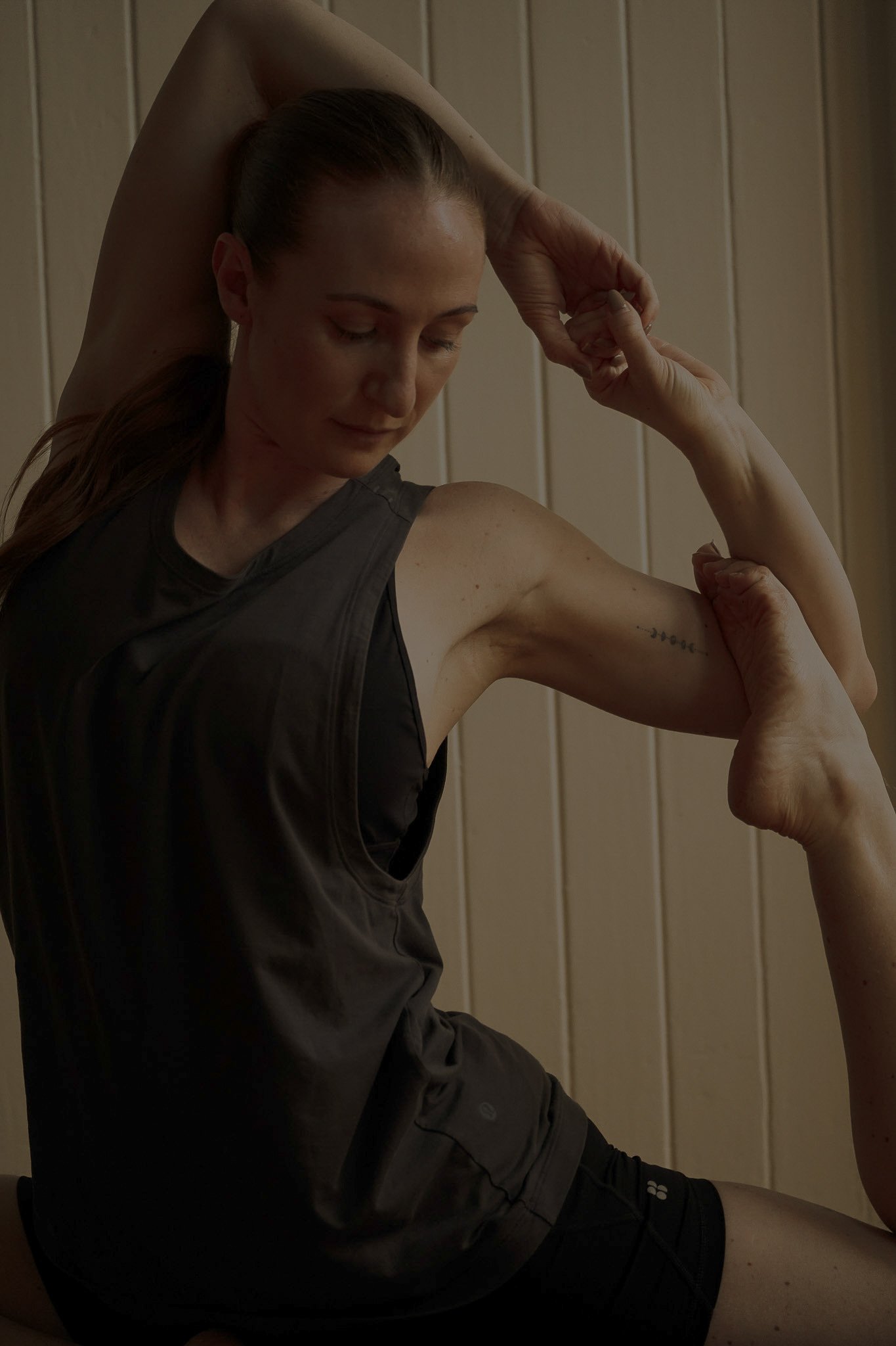 A woman with brown hair tied back practicing yoga, with her left arm bent and behind her head, and her right arm reaching to hold her left elbow, against a wooden wall.