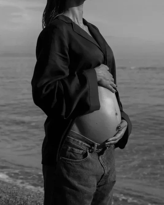 A pregnant woman standing on a beach, wearing a dark jacket and jeans, holding her belly with one hand.