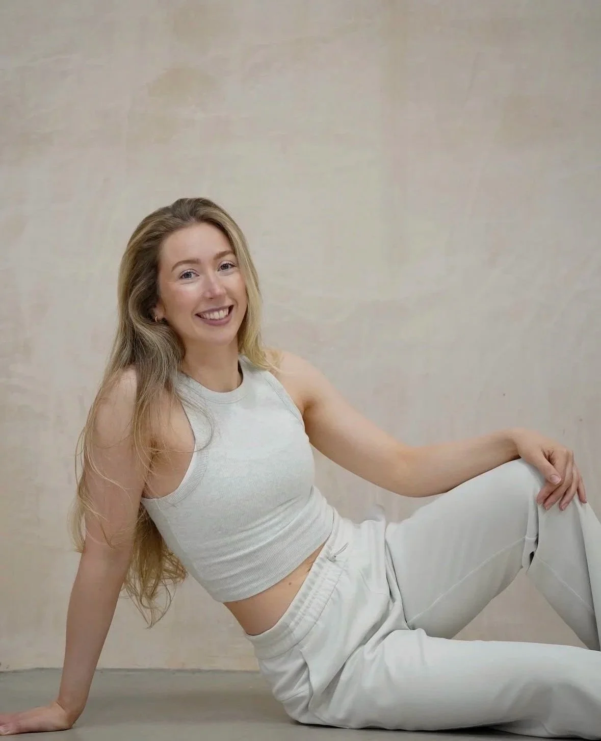 A young woman with long blonde hair, wearing a white sleeveless top and white pants, smiling and sitting on the floor against a plain wall.
