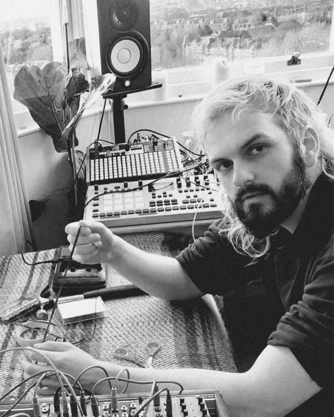A man with long, wavy, light-colored hair and a beard, sitting at a table with electronic music equipment, looking at the camera in a room with a window showing buildings outside.