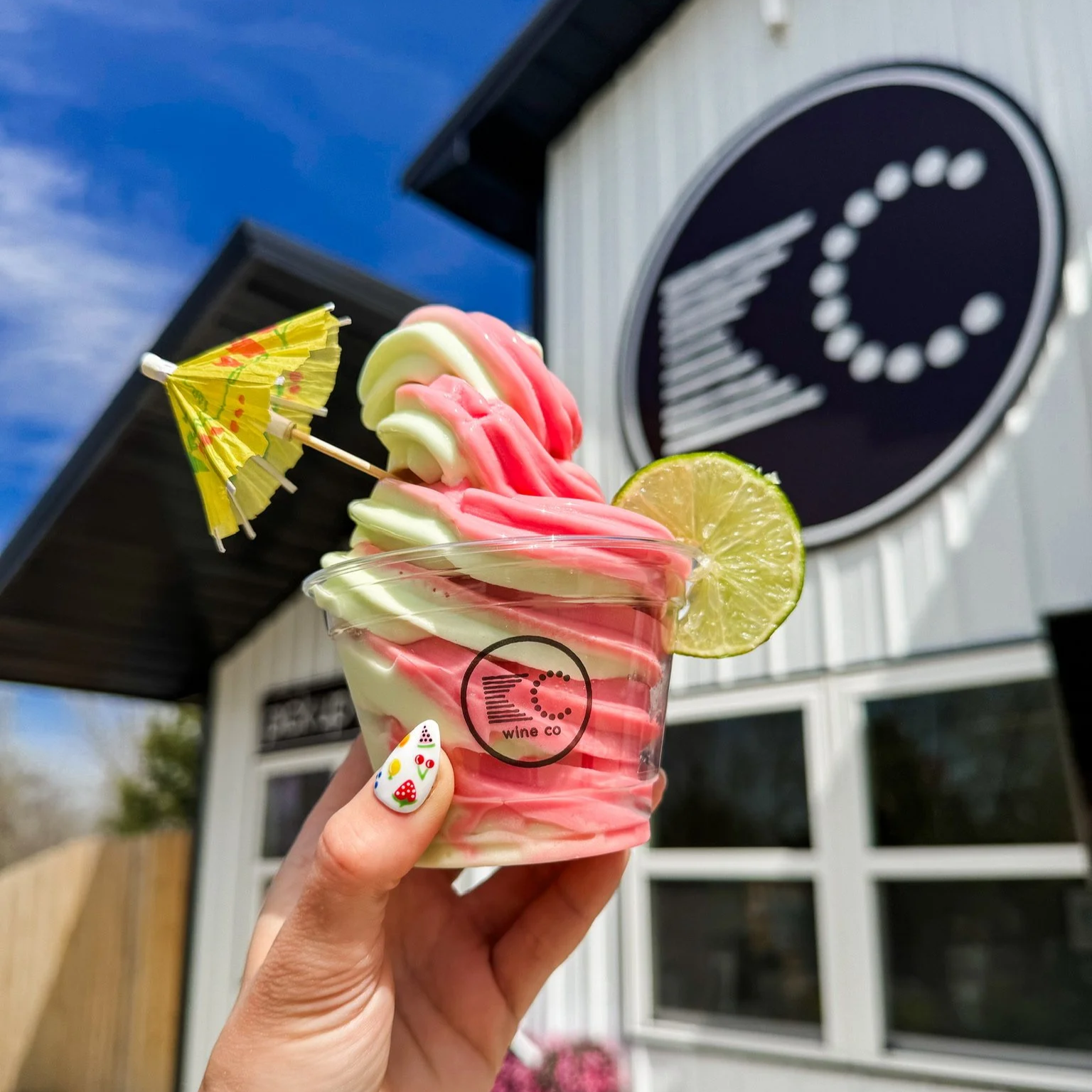 Hand holding up a soft serve twisted margarita with strawberry and lime flavors, topped with a lime slice and mini umbrella.