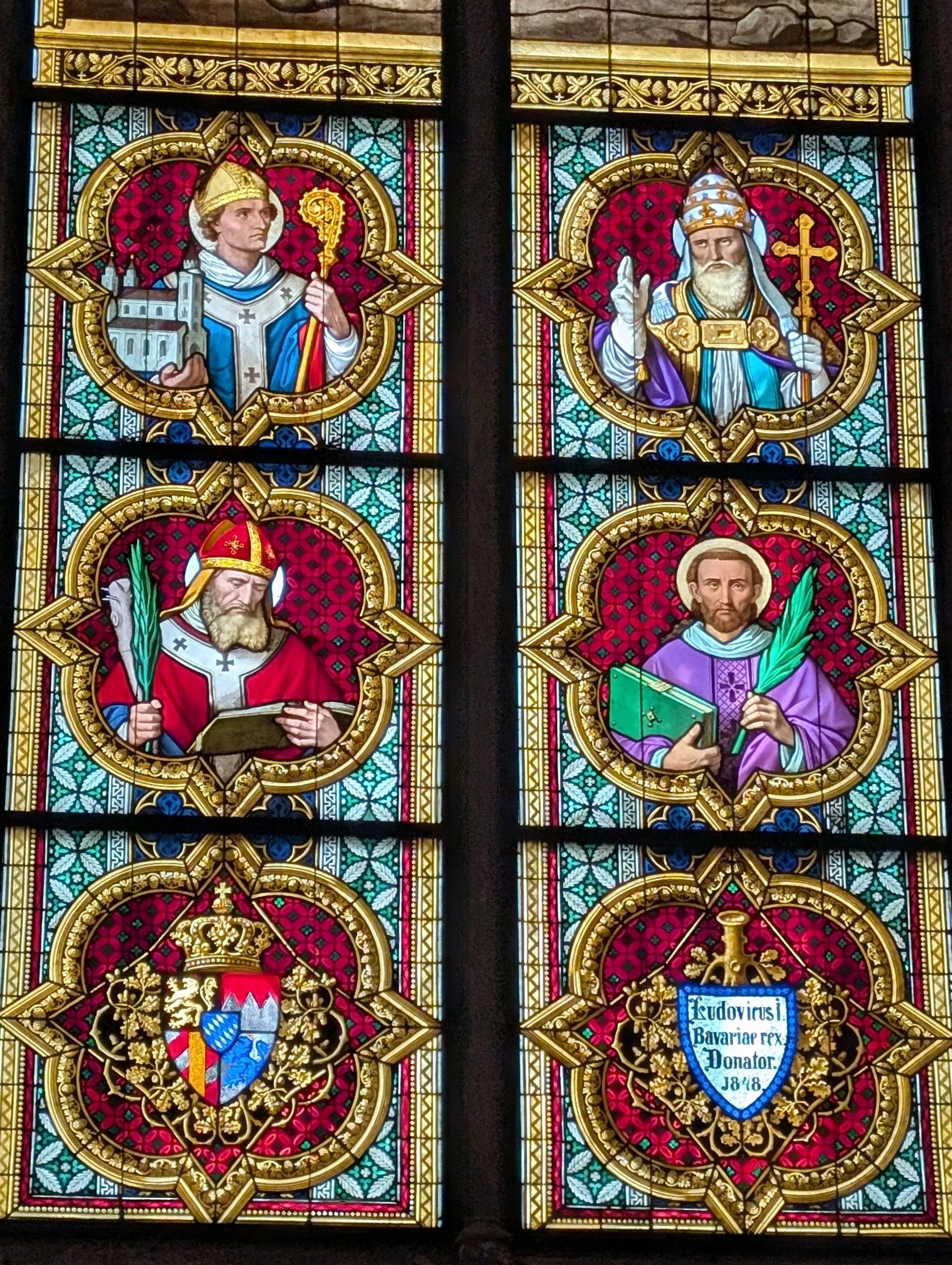 Stained glass window featuring portraits of religious and historical figures, with ornate gold and colorful backgrounds.