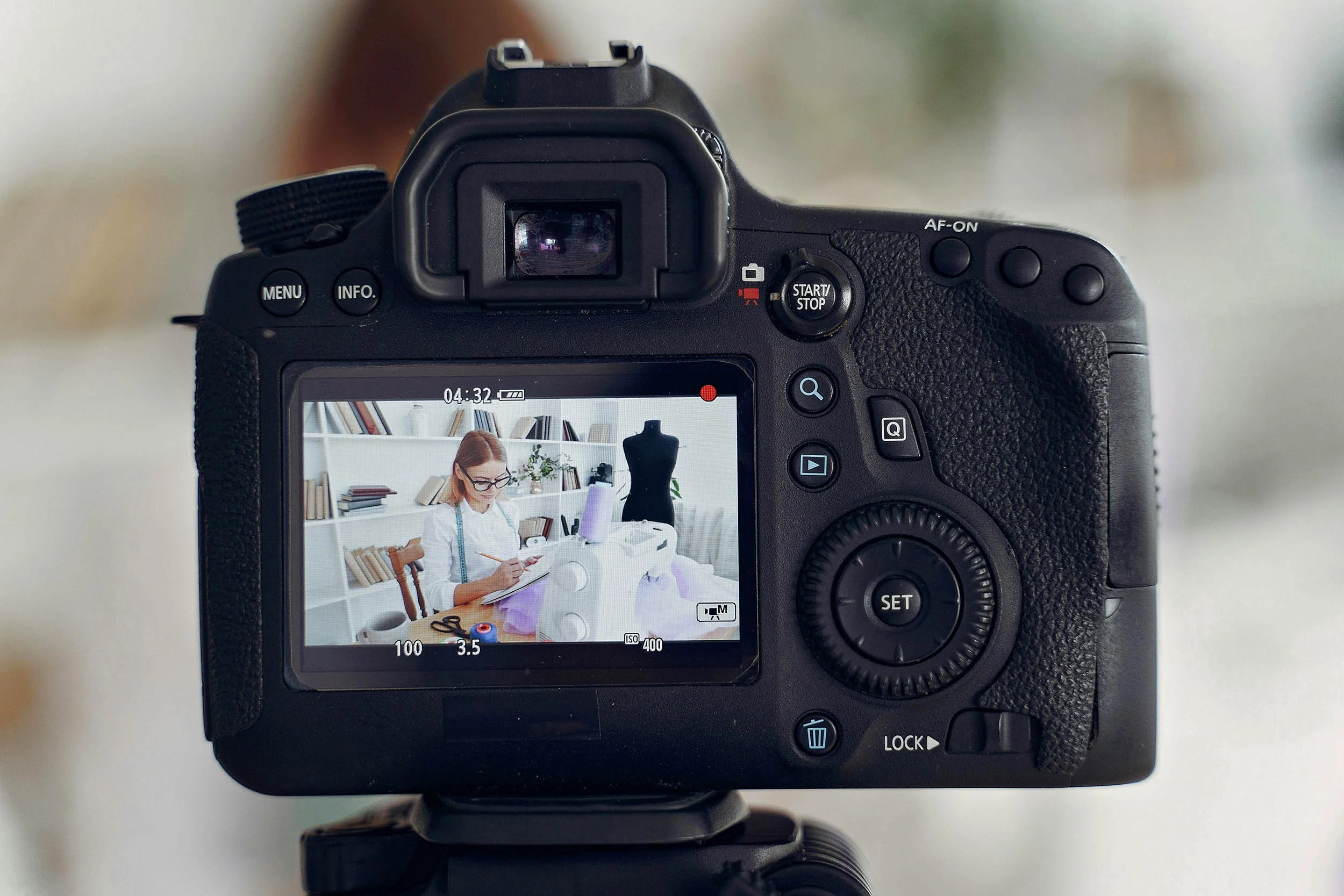 View of a professional digital camera capturing a woman working on a sewing project in a well-lit, organized room with shelves and a dress form in the background.