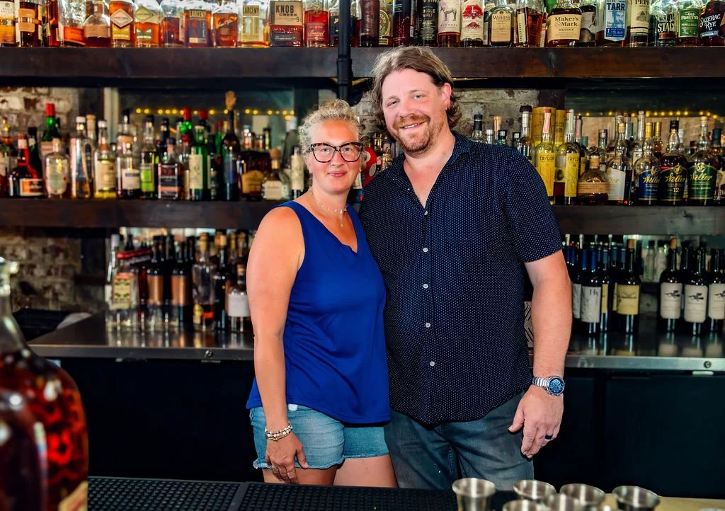 A woman and a man standing behind a bar counter, smiling, with shelves of various liquor bottles behind them in a bar or pub setting.