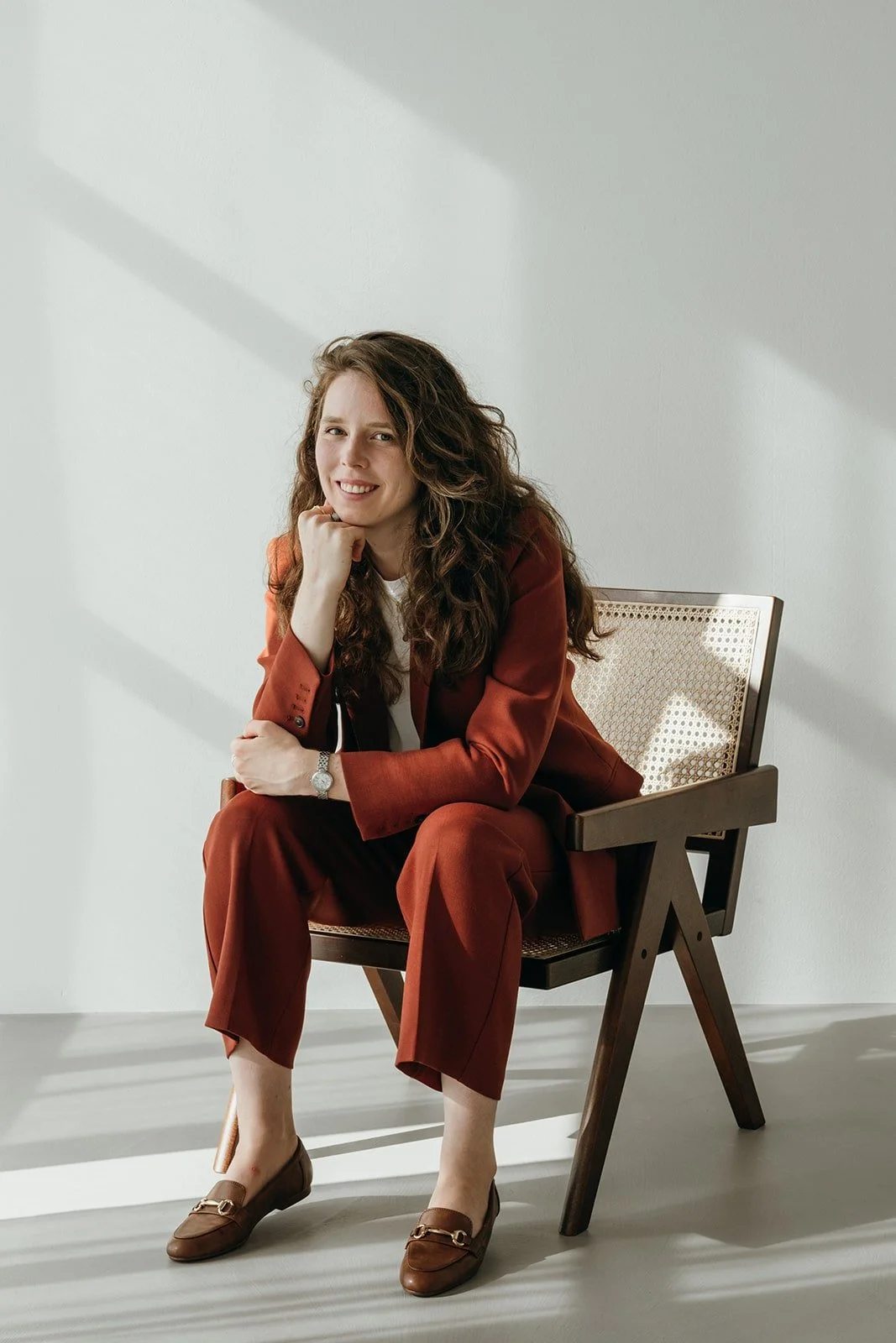 A woman with curly brown hair sitting on a wooden chair in a white room with sunlight coming through blinds, wearing a rust-colored pantsuit, white shirt, and loafers, smiling and resting her chin on her hand.