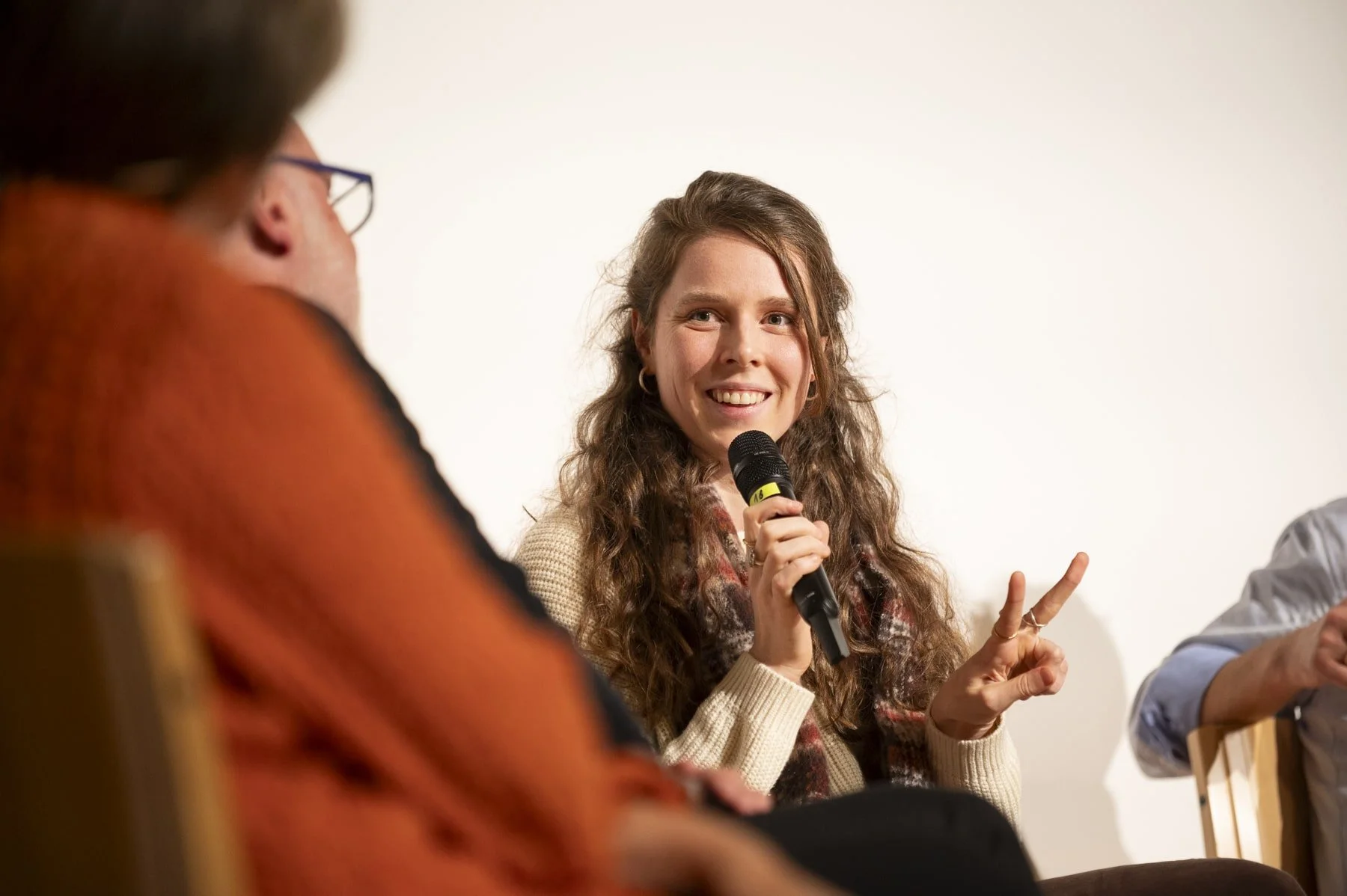 A woman with long curly hair smiling and holding a microphone, making a peace sign with her right hand during a panel discussion.
