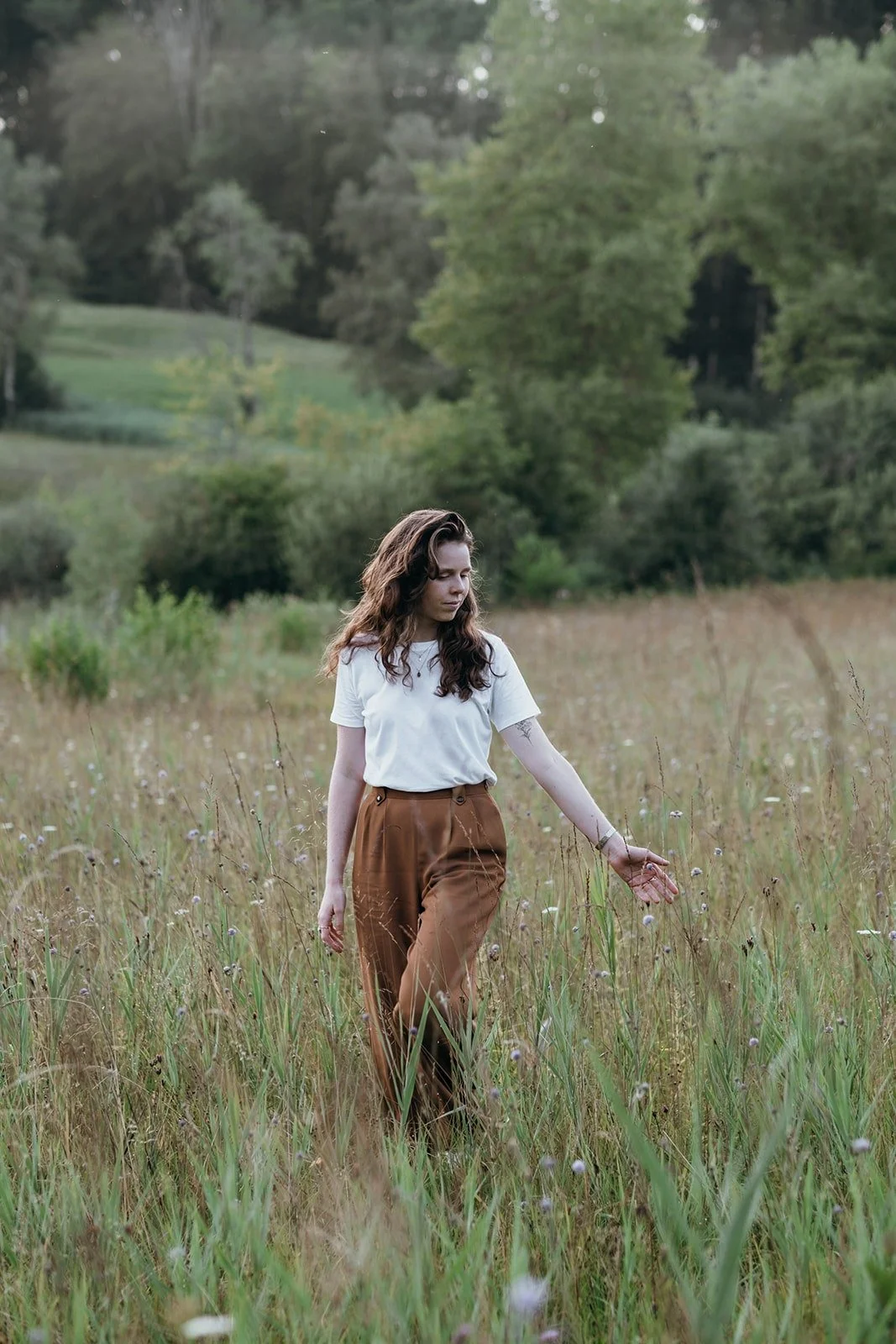 A woman with long brown hair wearing a white t-shirt and brown pants walks through a grassy meadow with wildflowers, surrounded by green trees and hills in the background.