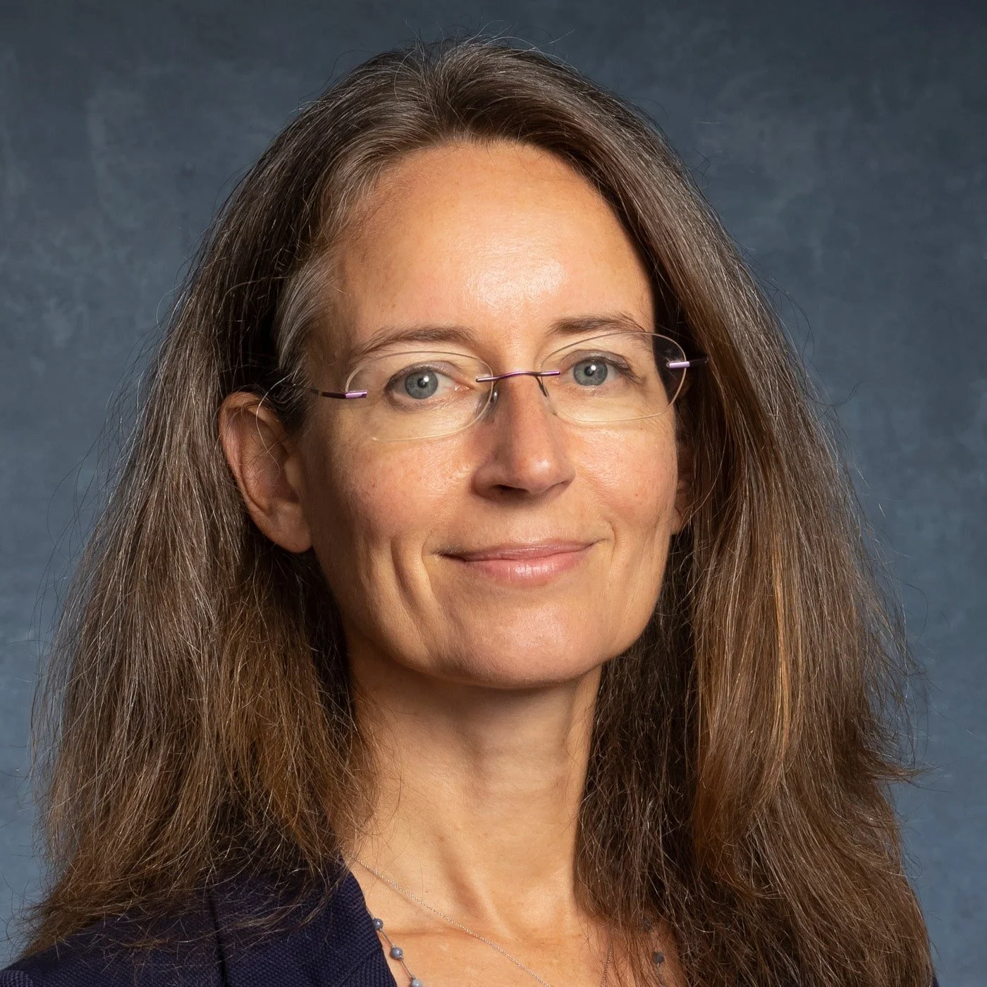 A middle-aged woman with long brown hair, wearing glasses, a necklace, and a dark blazer, smiling at the camera against a blue-gray background.
