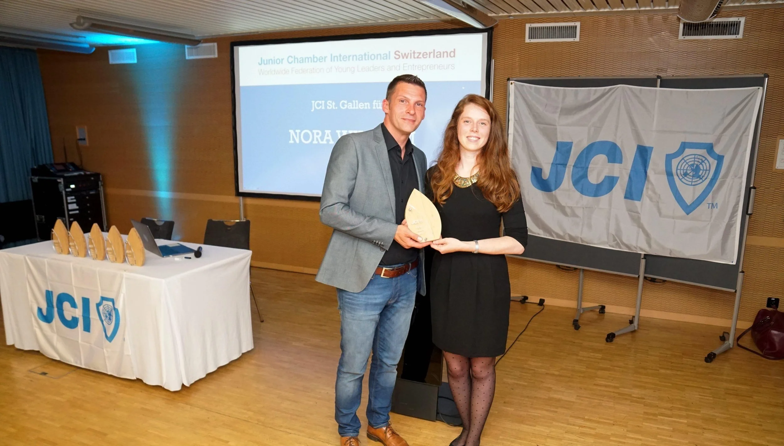 A man and woman holding a wooden award at a JCI event, with a large JCI banner and a screen displaying event information in the background.