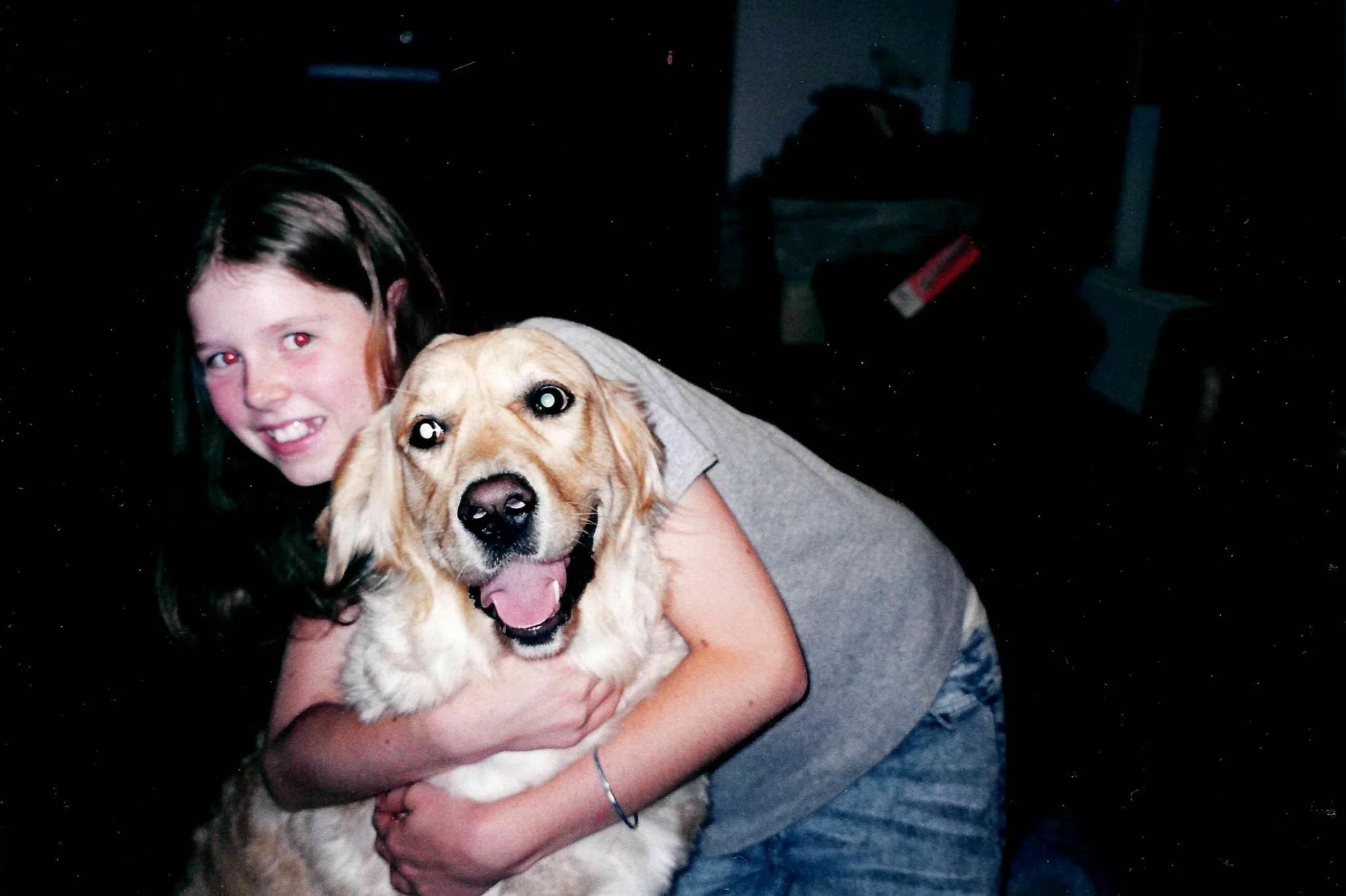A young girl with dark hair hugging a happy golden retriever dog inside a dimly lit room.