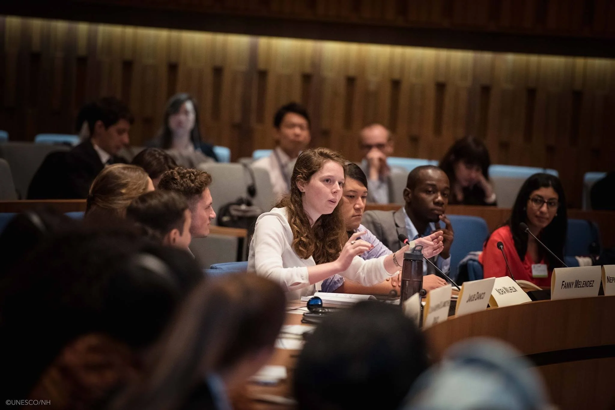 People attending a conference or meeting, sitting at tables with nameplates, microphones, and notebooks in a wooden-paneled room.