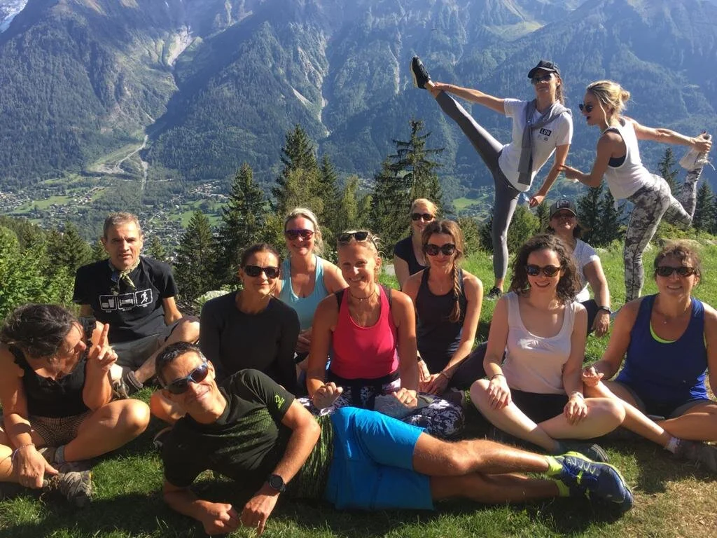 A group of people posing on a grassy hillside with a mountainous landscape in the background. Two individuals are performing yoga poses. The scene is sunny, and all are dressed in casual outdoor attire.