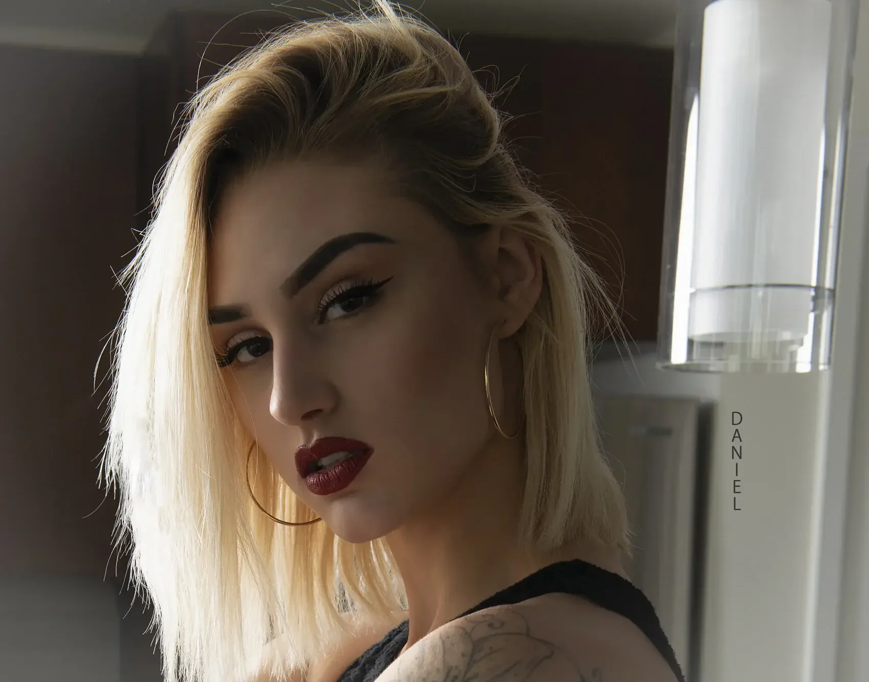 A woman with platinum blonde hair, winged eyeliner, dark red lipstick, and hoop earrings, looking over her shoulder at the camera, standing in front of a kitchen appliance.